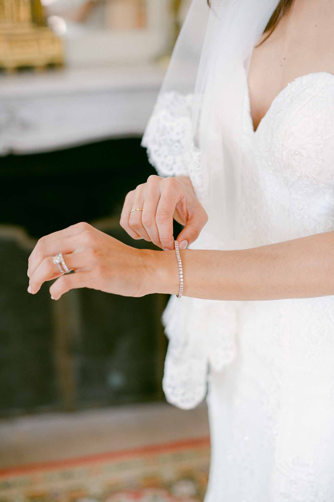 Bride fastening diamond tennis bracelet over white lace gown with engagement ring and wedding band visible