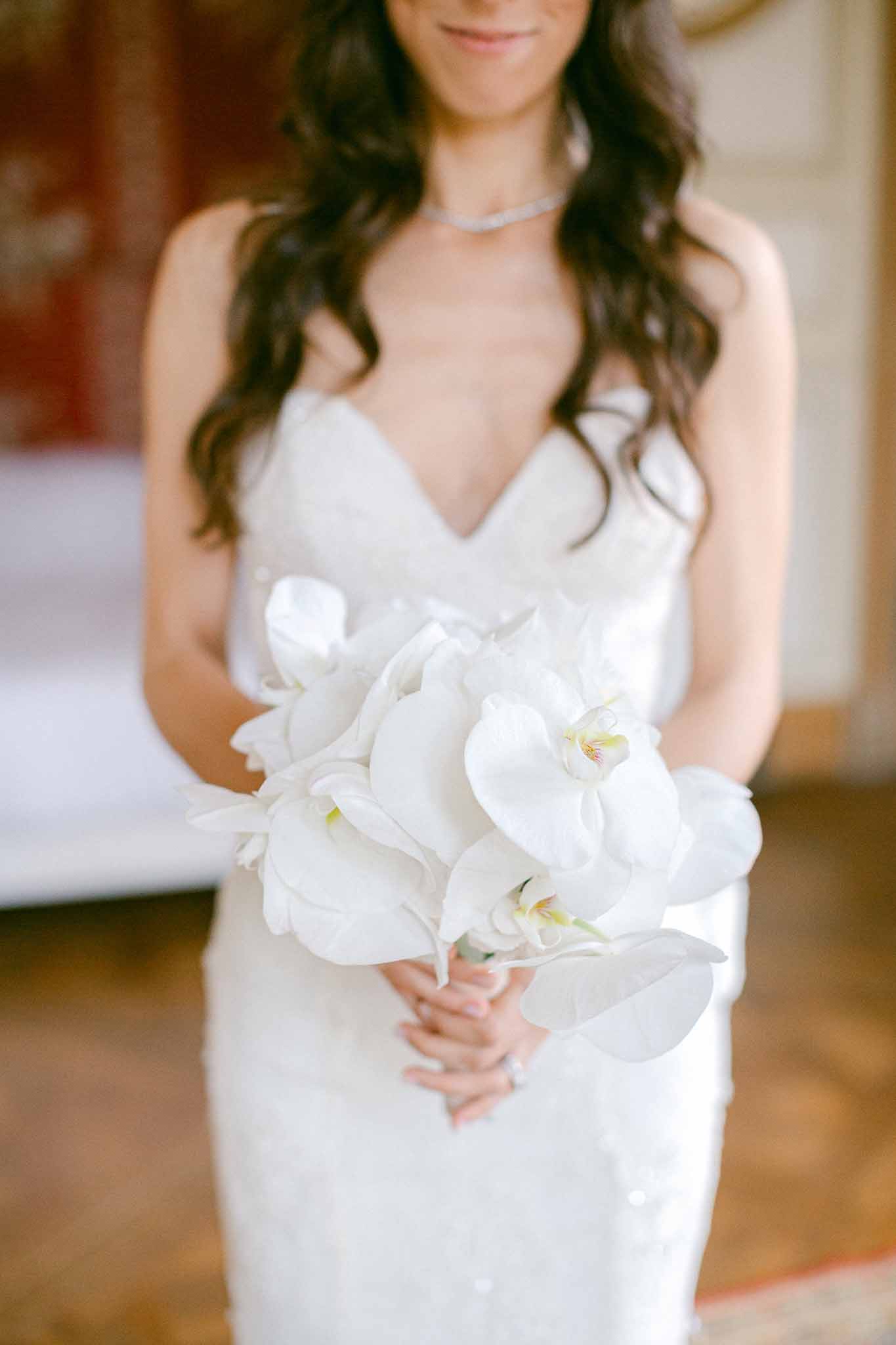 Close-up of bride holding rounded white phalaenopsis orchid bouquet against ivory gown