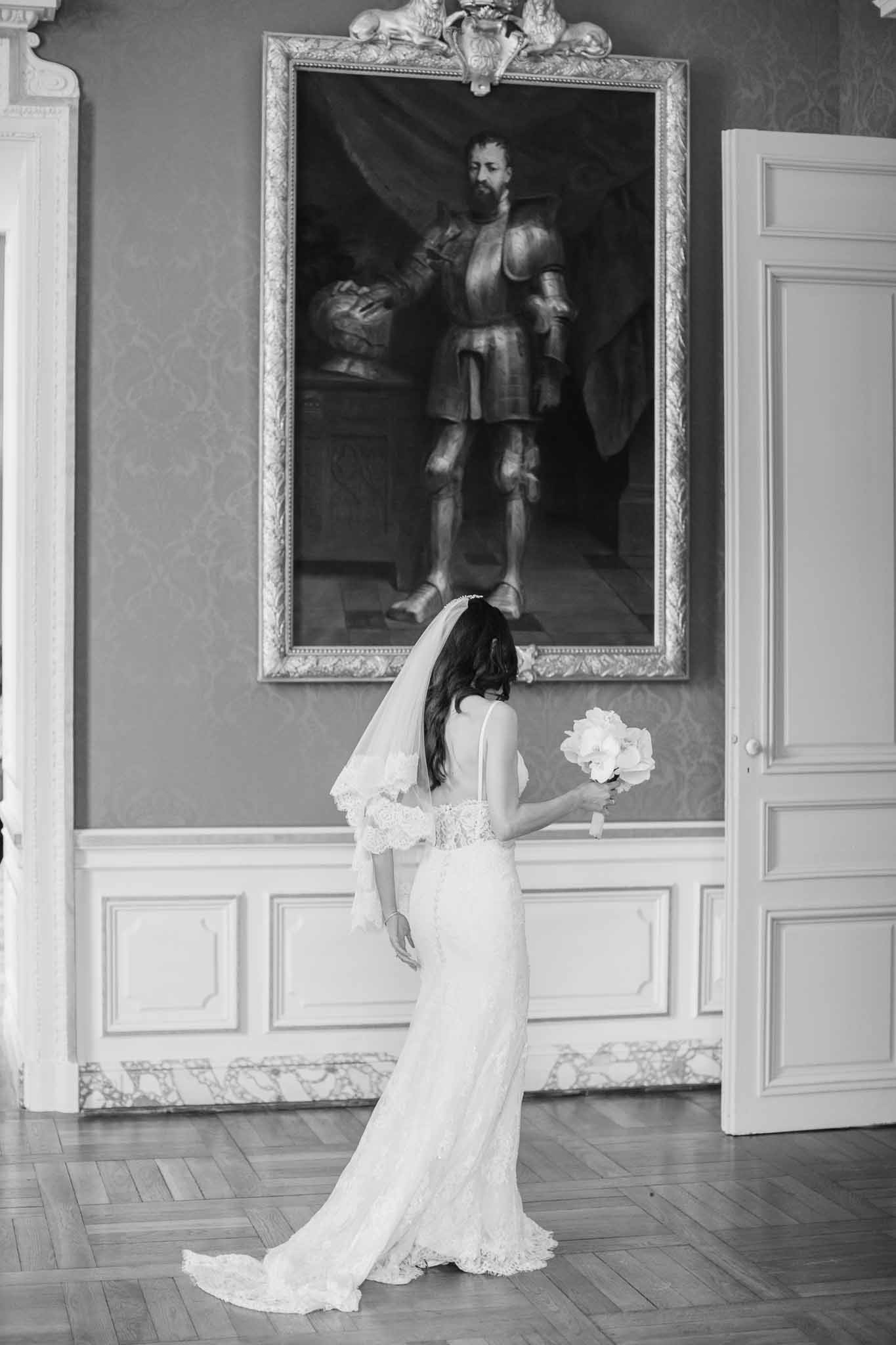 Black and white portrait of bride from behind facing gilt-framed painting in chateau room with damask walls
