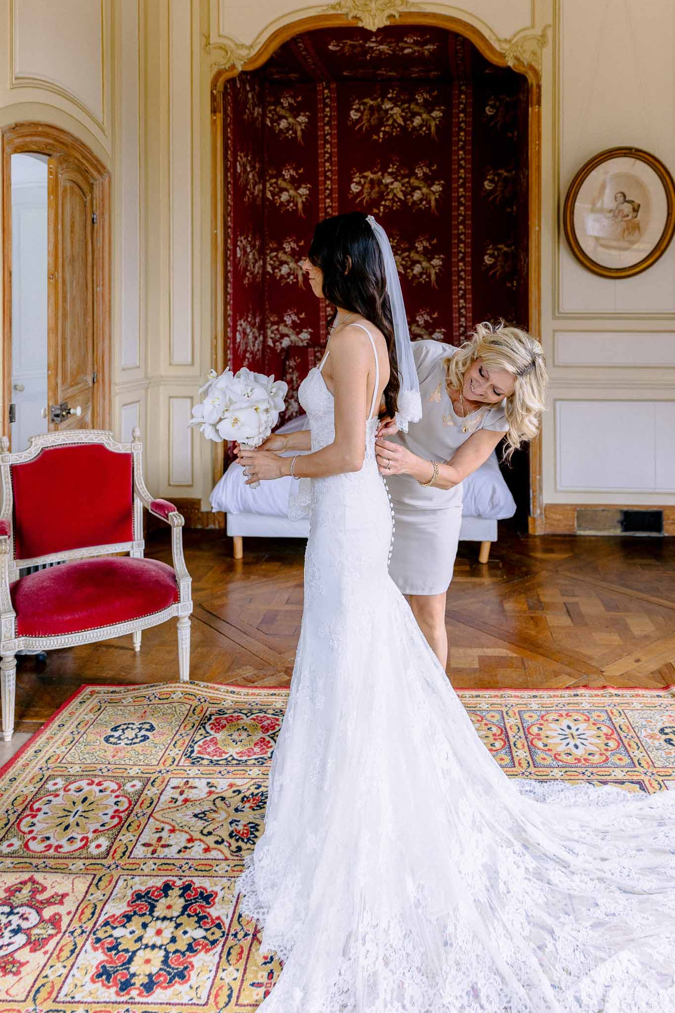 Assistant fastening bride's lace mermaid gown and cathedral train in boiserie room with red Persian rug