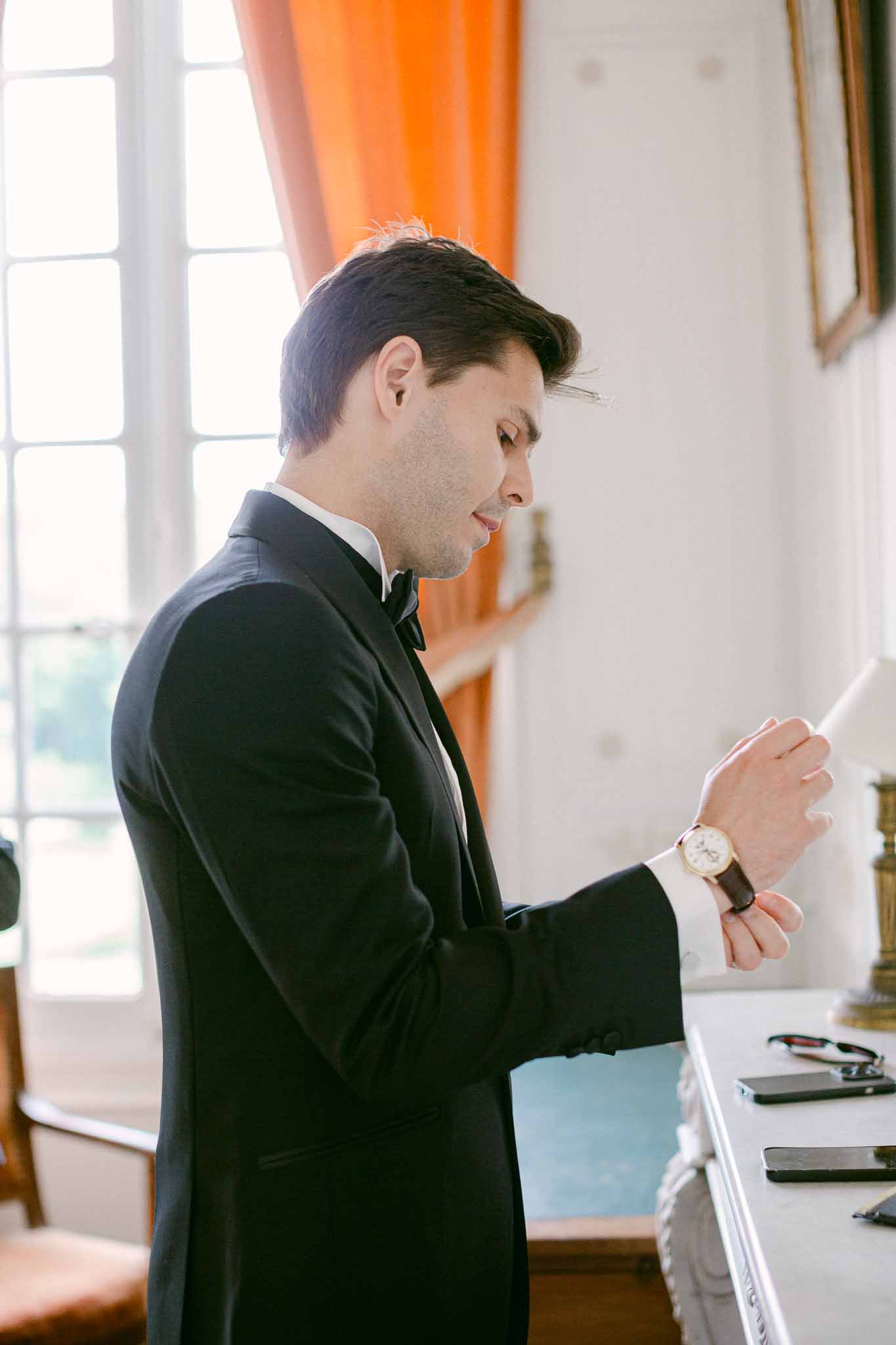 Groom in black tuxedo reading letter beside tall French window with orange curtains in white panelled chateau room