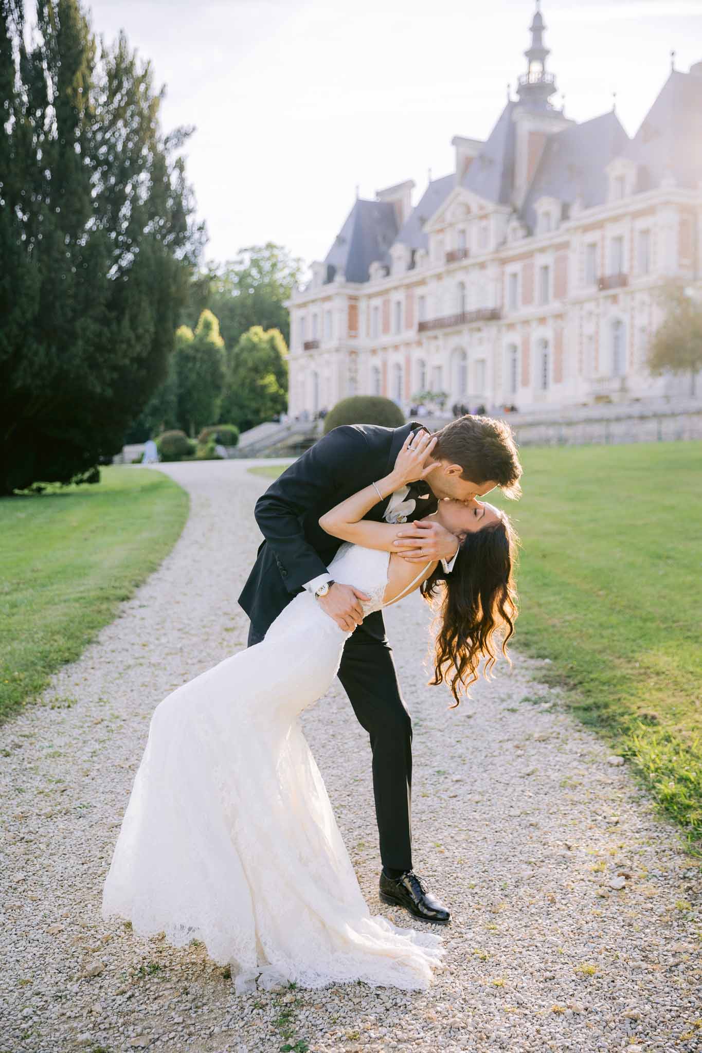 Groom dipping and kissing bride in ivory lace mermaid gown on gravel drive in front of French chateau at golden hour