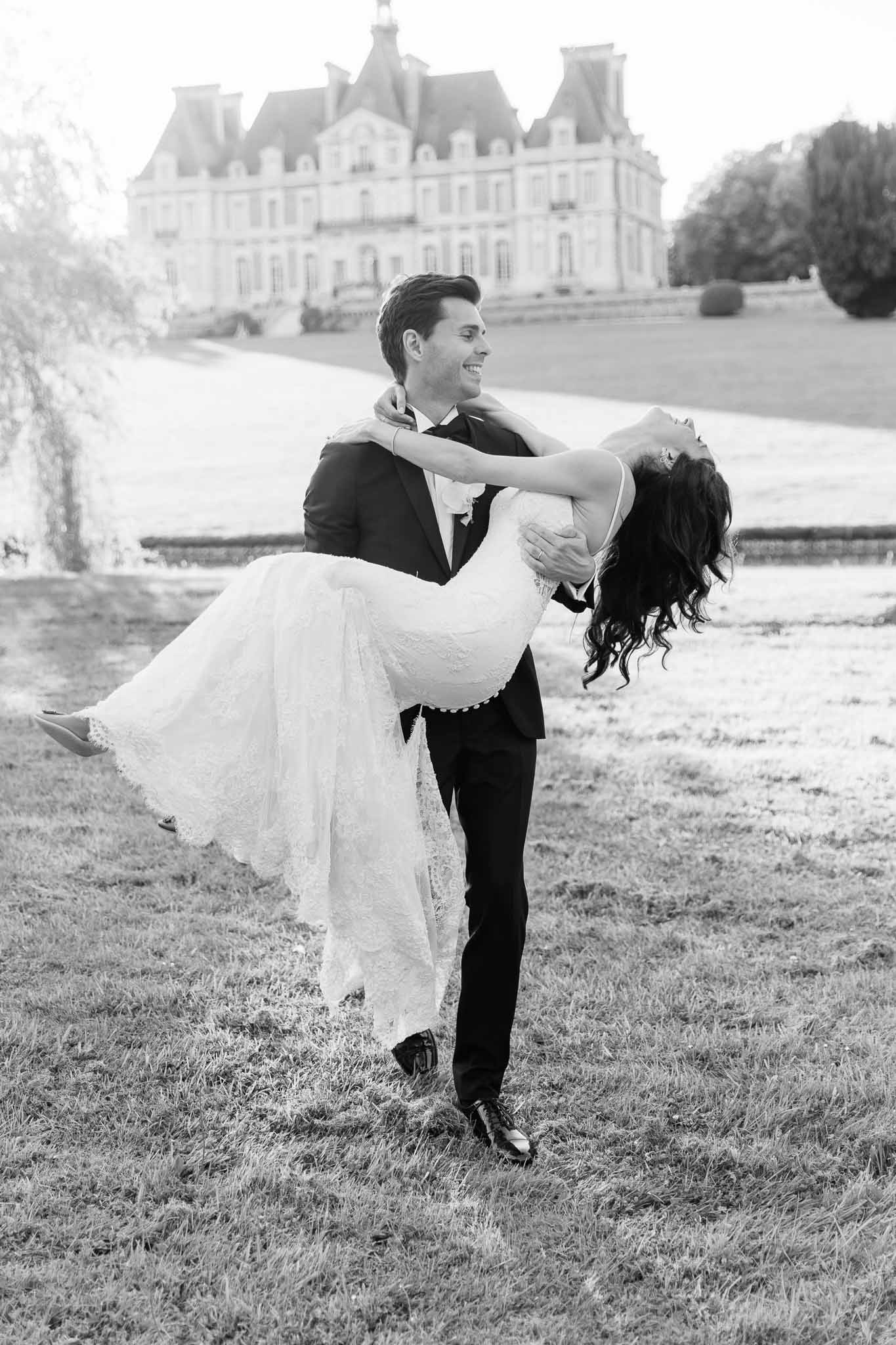 Black and white groom lifting bride in lace gown on chateau grounds with classical facade and gravel driveway behind