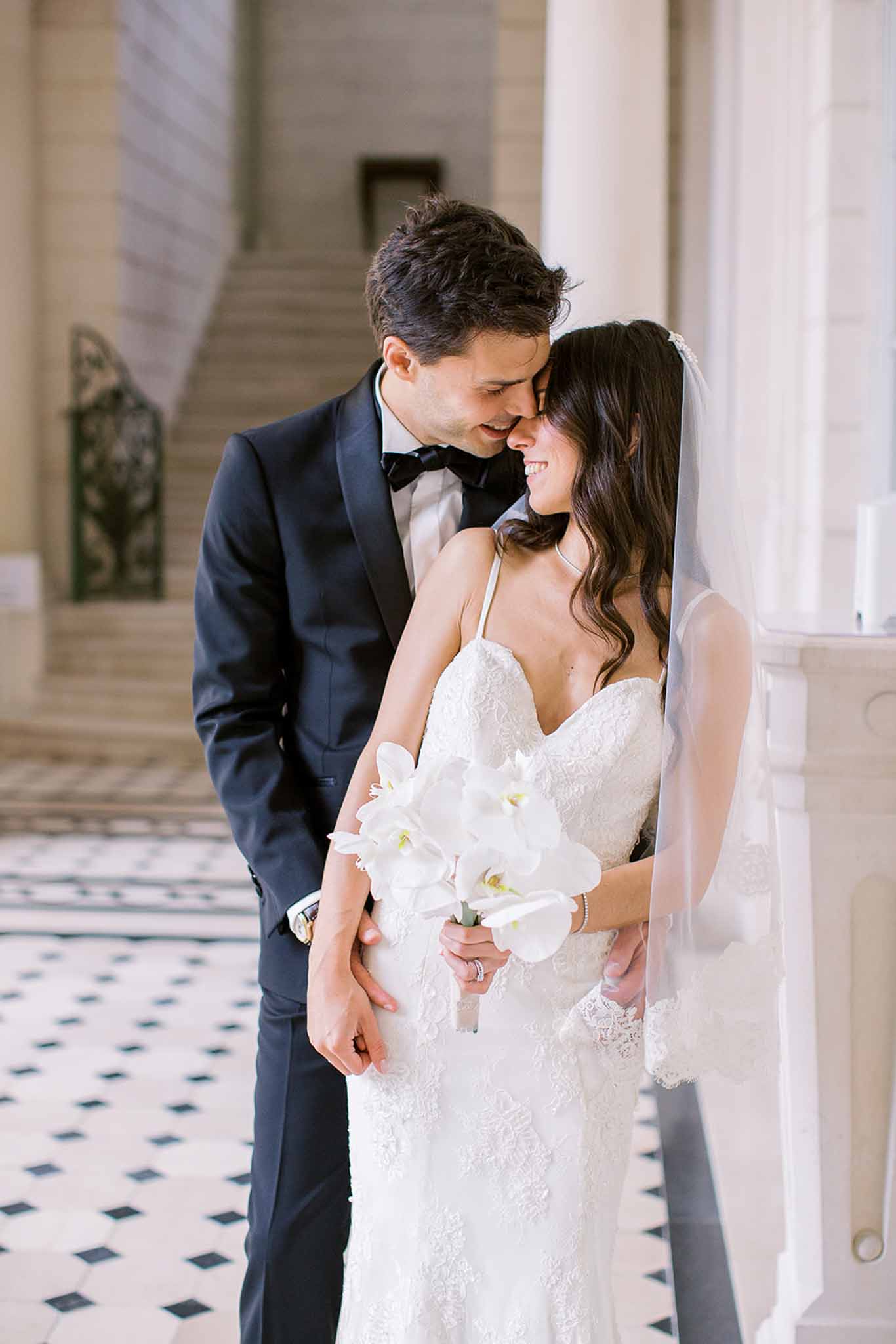 Bride in ivory lace gown with beaded veil and groom in navy suit touching foreheads on marble floor