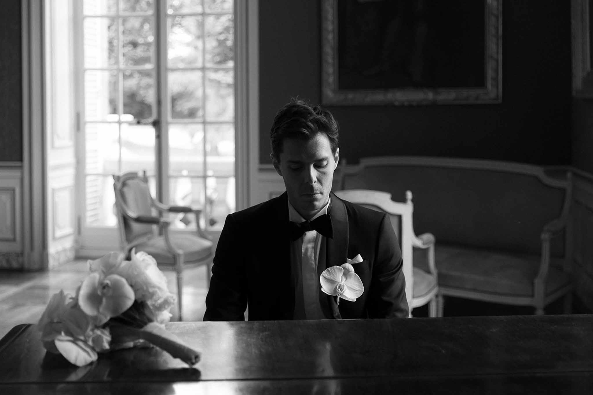 Black and white of groom in tuxedo seated at table with orchid bouquet in chateau salon with gilt painting and Louis chairs