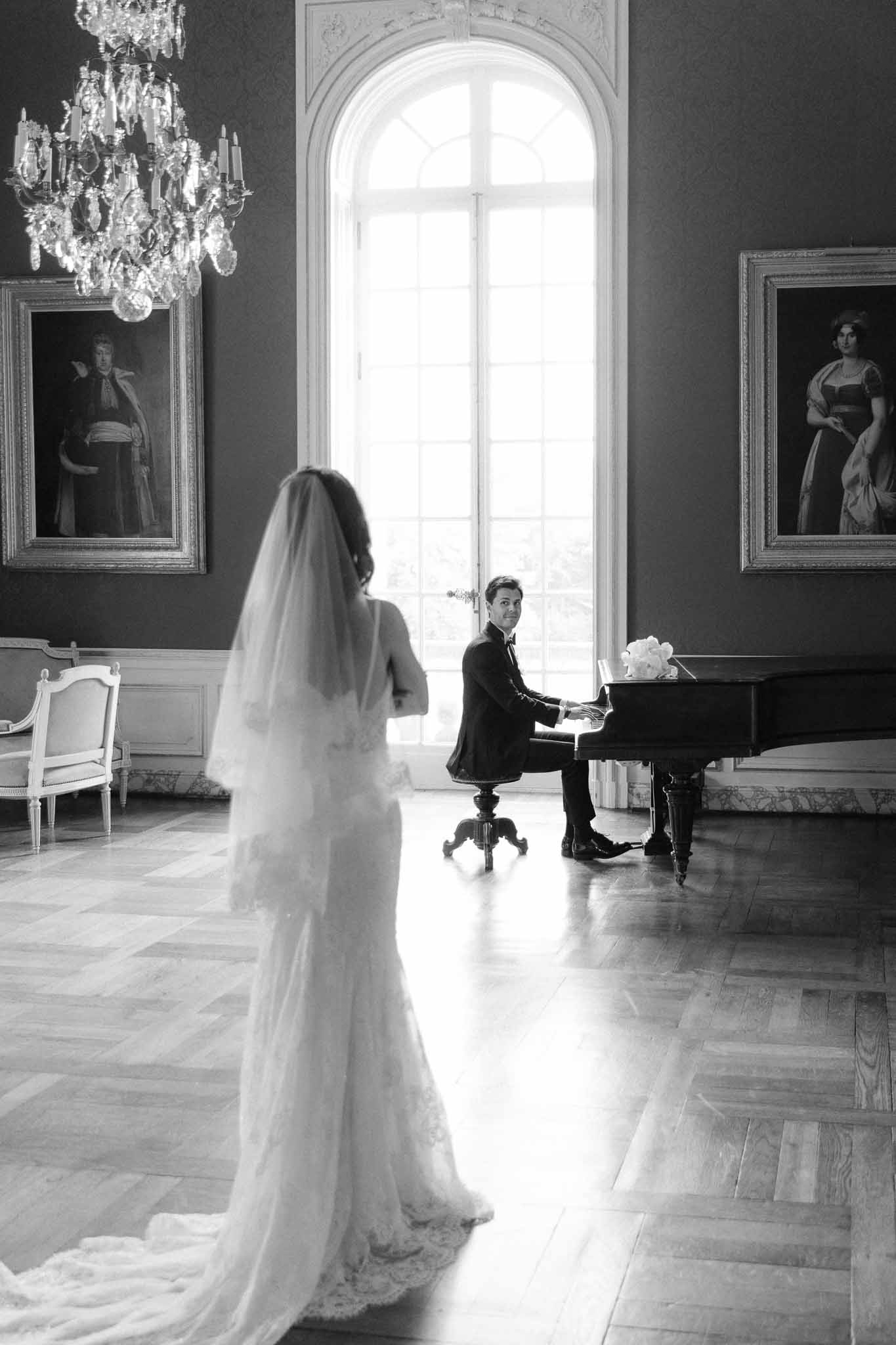 Black-and-white portrait of groom at grand piano and bride approaching in lace gown inside chateau salon