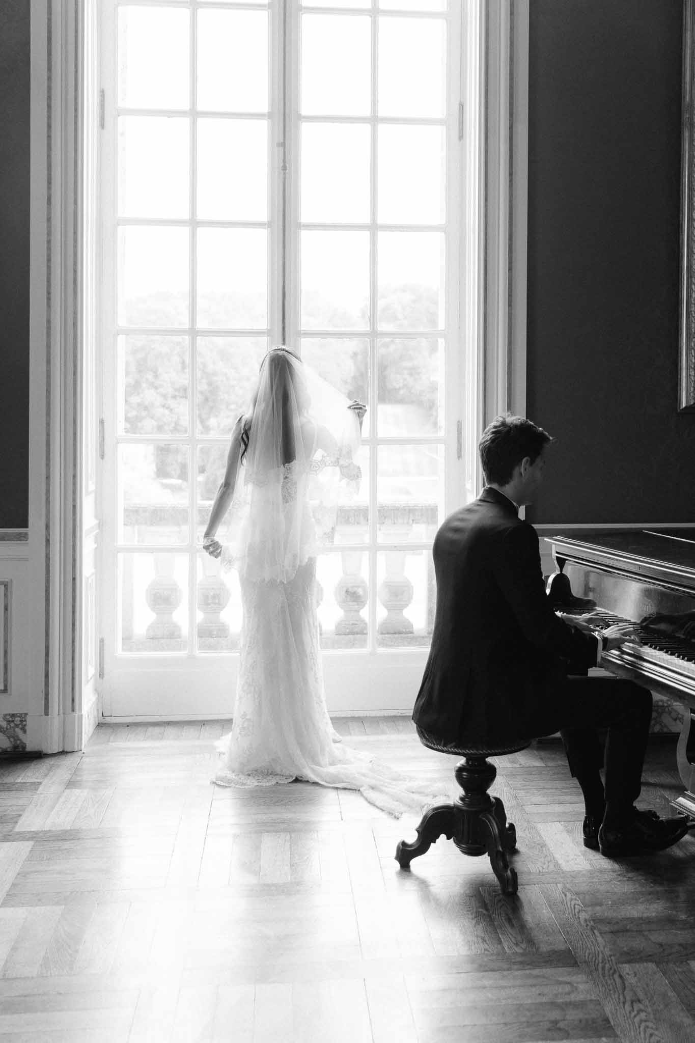 Black and white of groom at piano as bride stands in lace gown and veil at backlit French doors