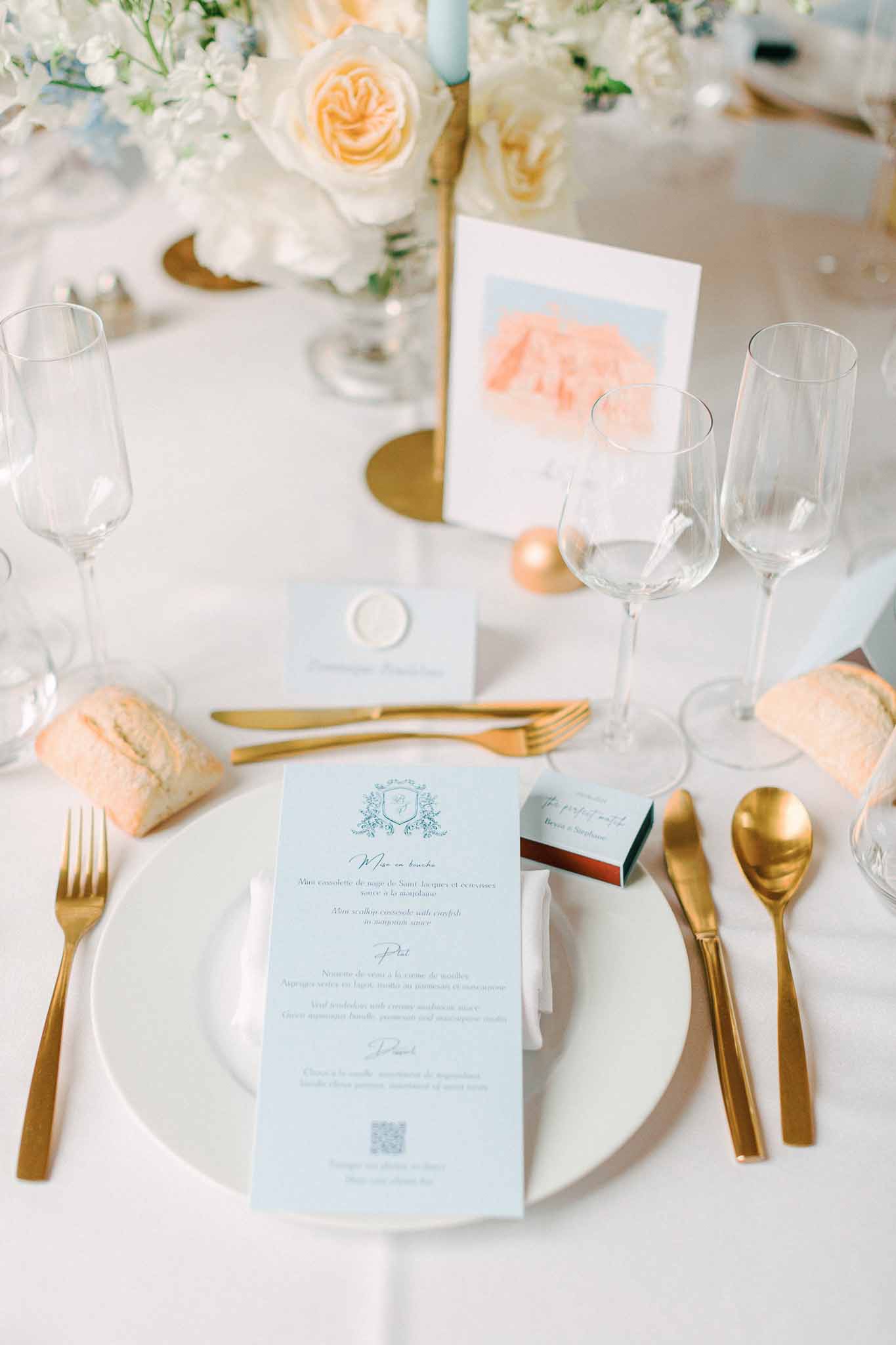 Place setting with gold flatware, pale blue menu card, ivory roses in gold vessel and blue taper candle