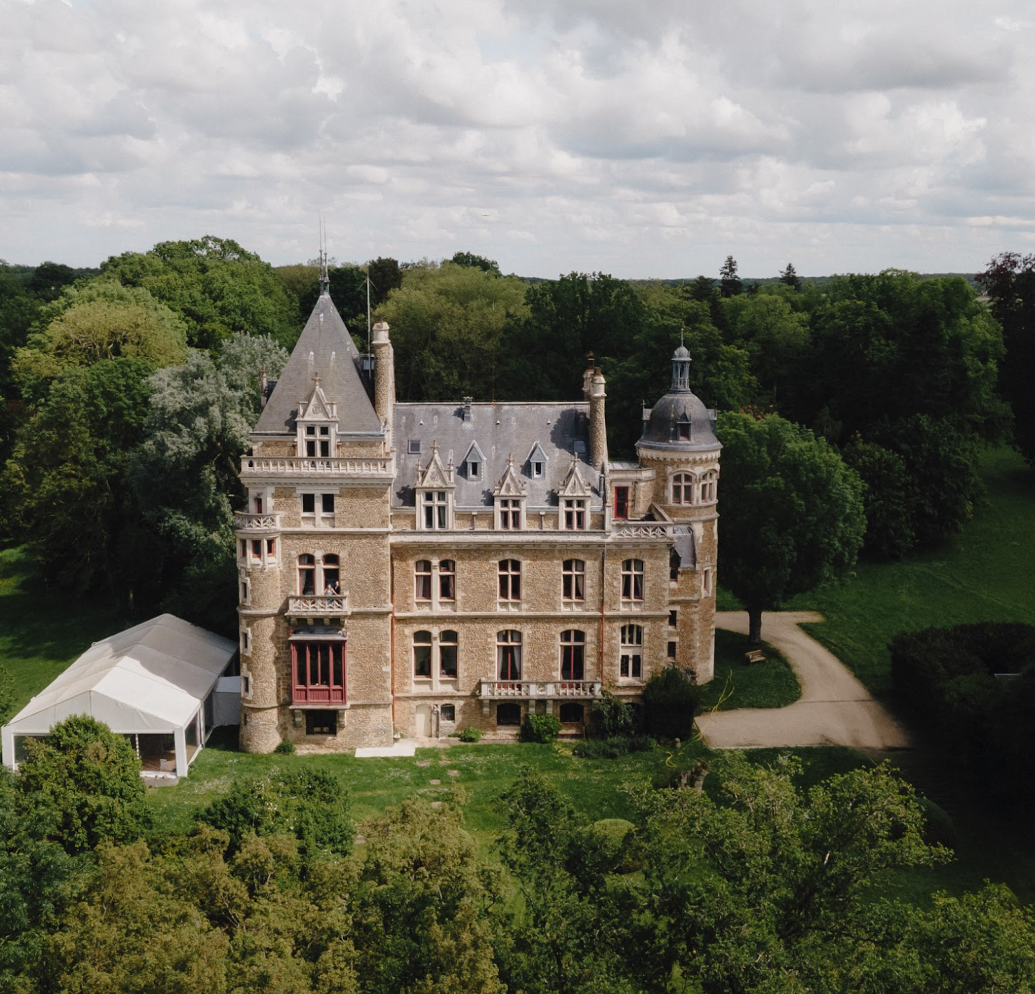 Aerial view of sandy-beige chateau with slate turrets red-trimmed windows white marquee and gravel driveway