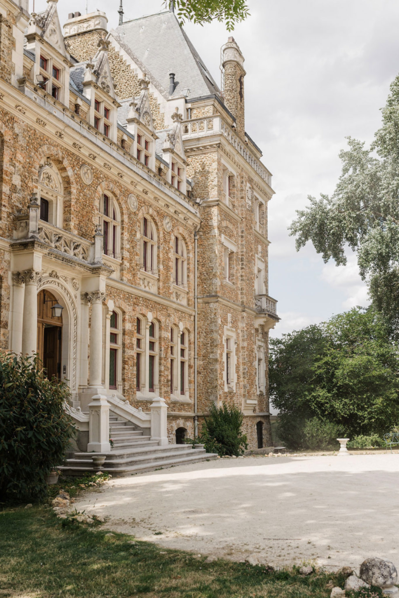 Exterior facade of French chateau with golden stone walls, slate-roofed corner tower, columned entrance portico and gravel...