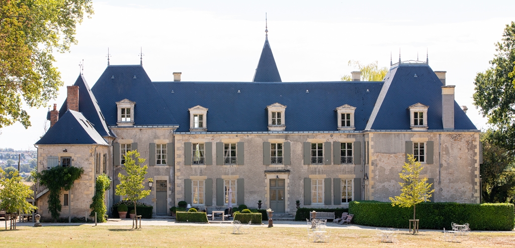 Cream stone chateau with dark slate mansard roof, pointed turrets, grey shutters, and topiary-lined terrace in daylight
