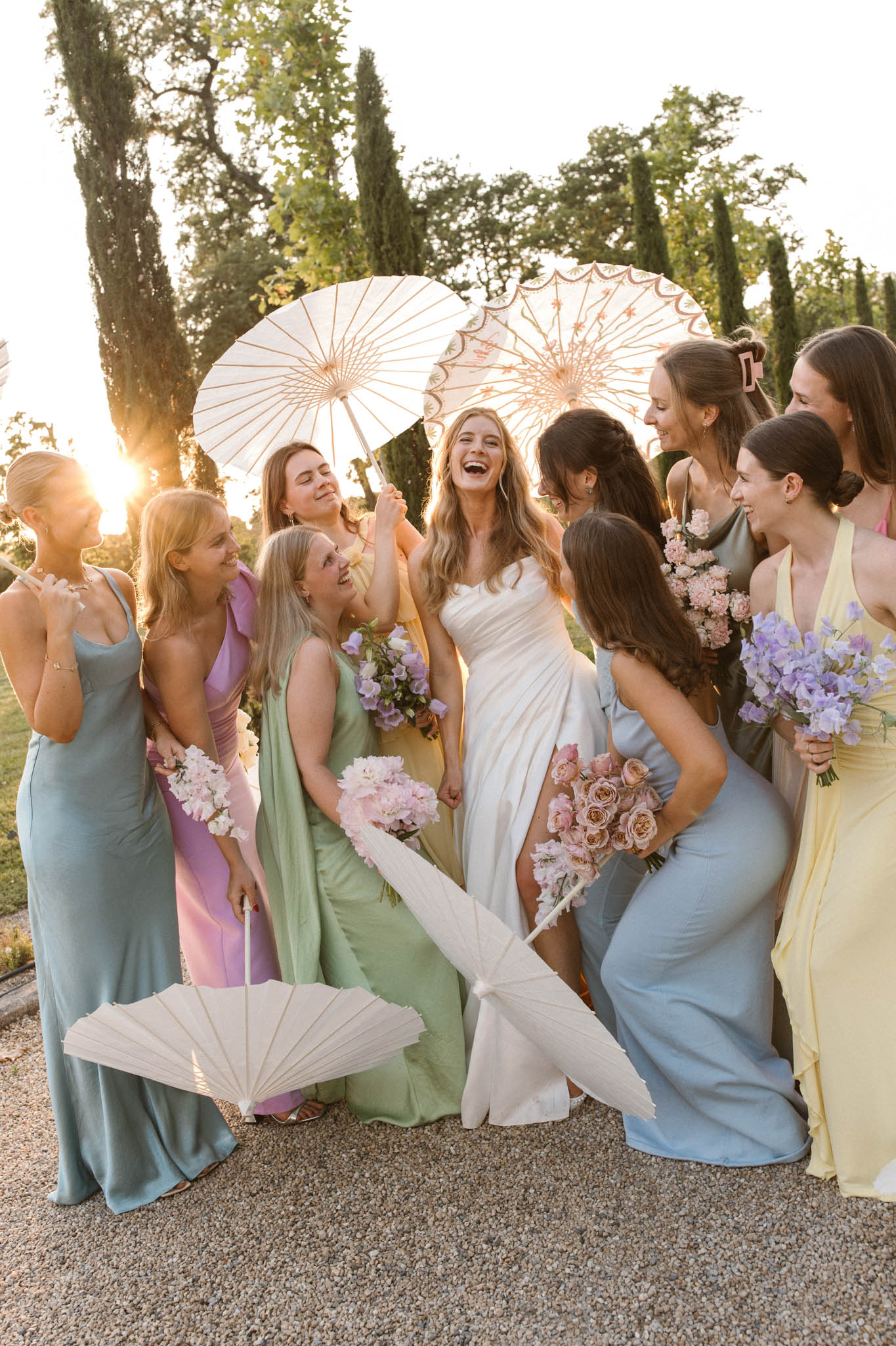 Bride with eight bridesmaids in mismatched pastel gowns holding varied bouquets at golden-hour on gravel path