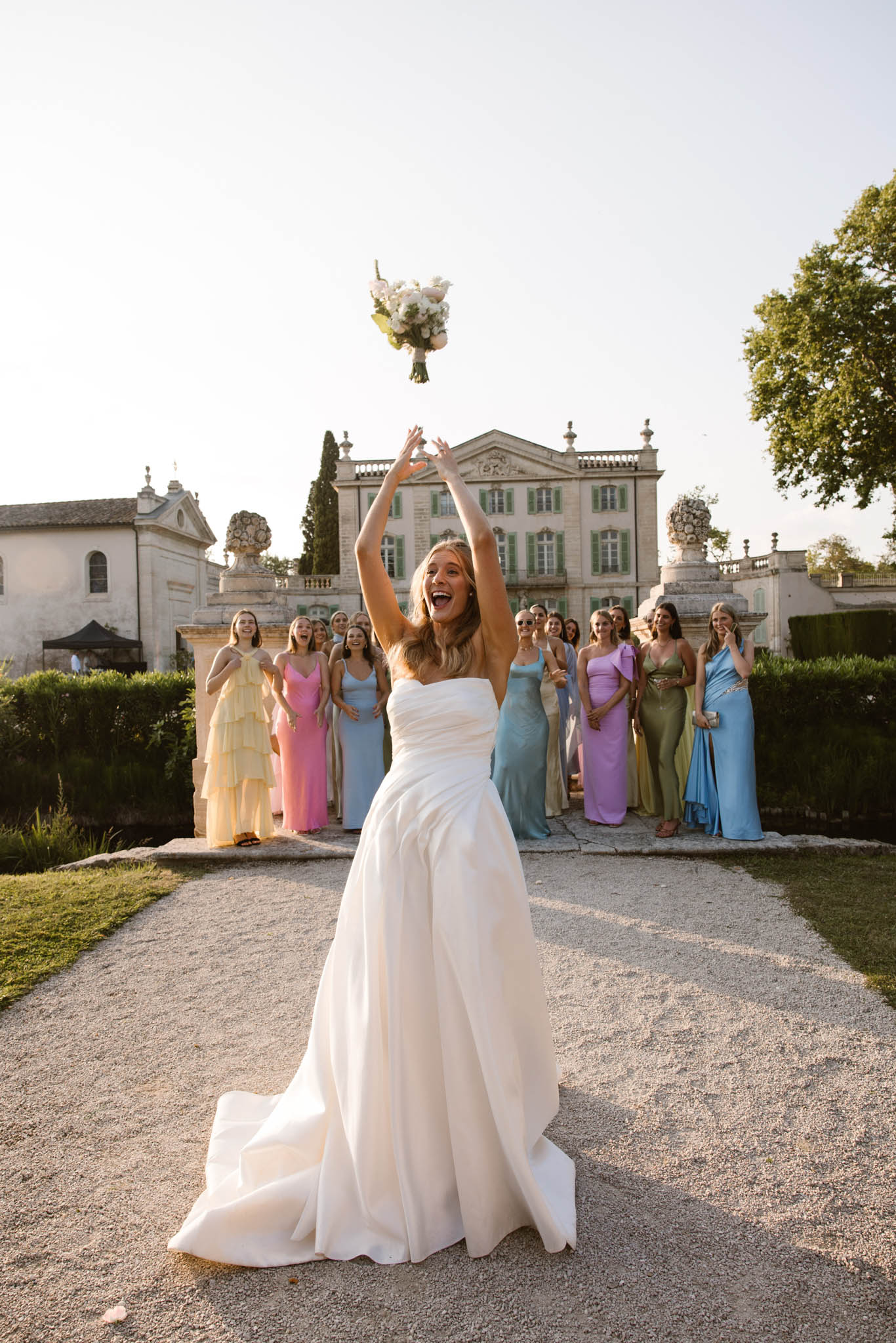 Bride tossing white peony bouquet with fifteen guests in colorful gowns before chateau at golden hour