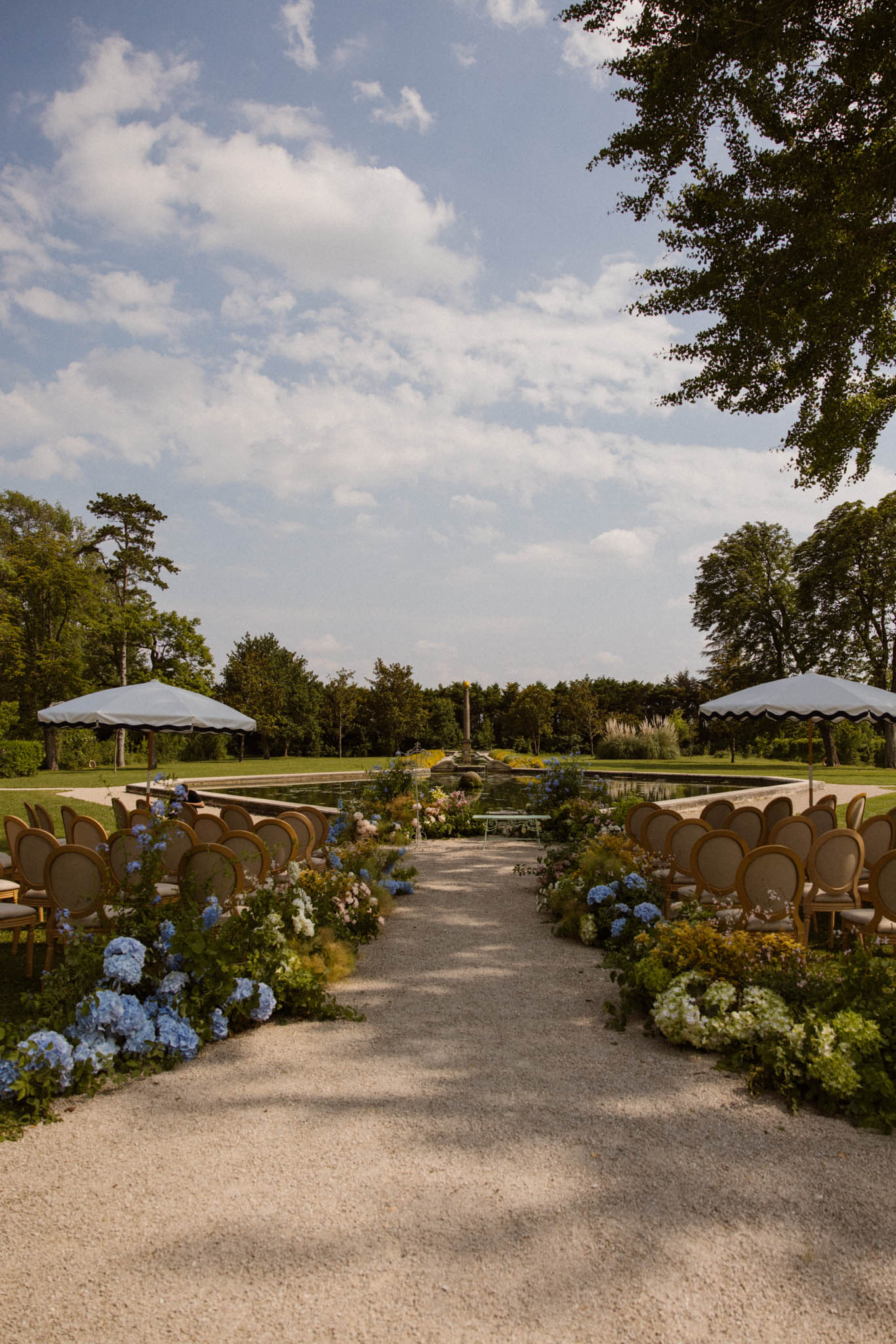 Gold Louis XVI chairs flanking aisle with blue hydrangea and blush borders before reflecting pool and white umbrellas