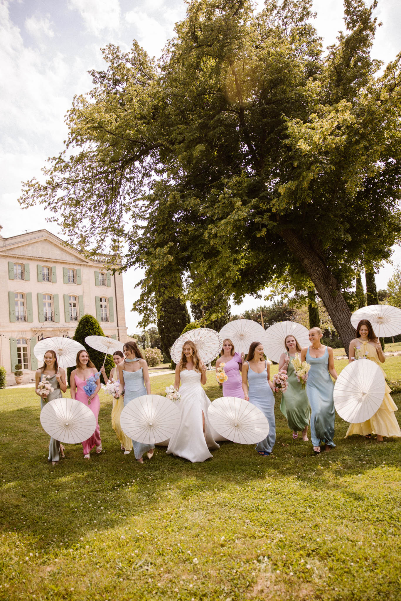 Bride and ten bridesmaids in mismatched pastel rainbow dresses with paper parasols crossing lawn