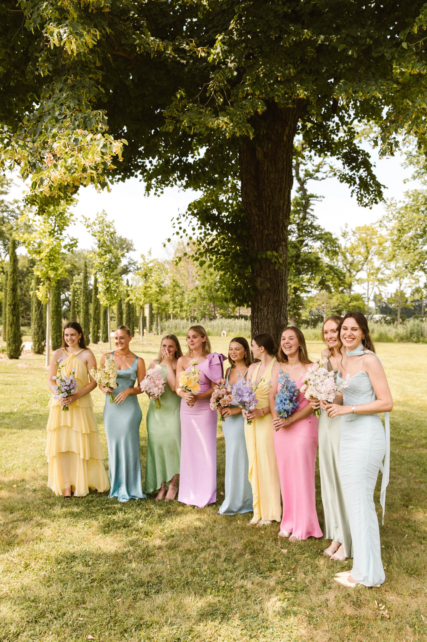 Nine bridesmaids in mismatched pastel rainbow dresses holding coordinating bouquets in park