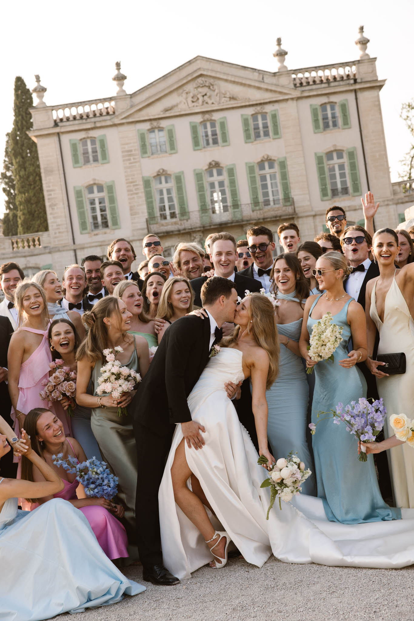 Groom dipping bride as 35 guests in pastel gowns and tuxedos cheer before stone chateau facade