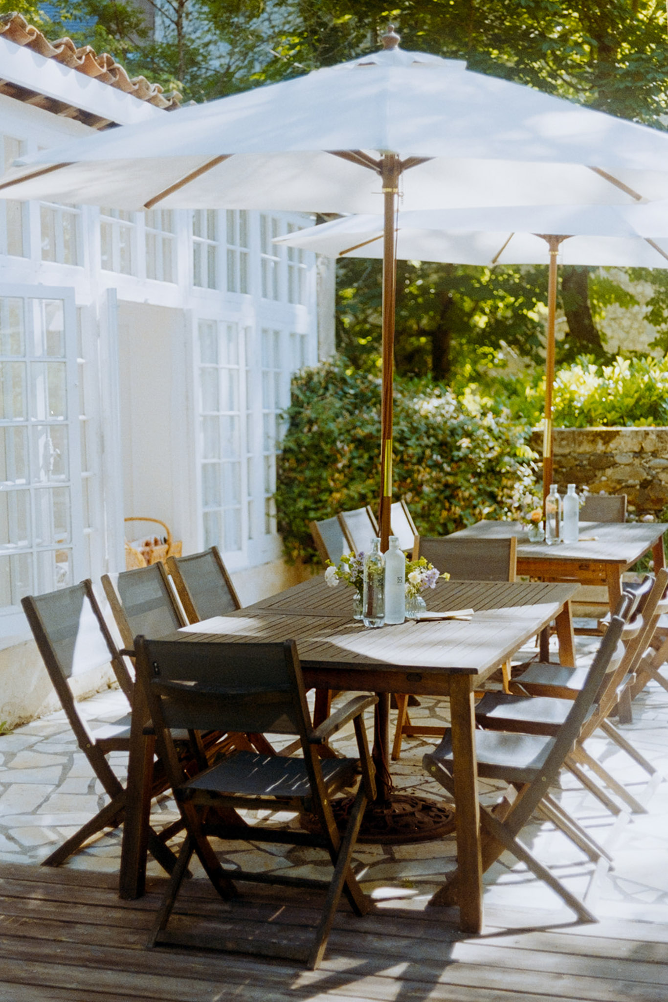 Outdoor terrace with two long wooden trestle tables, canvas chairs, white umbrellas, and simple bud vase florals