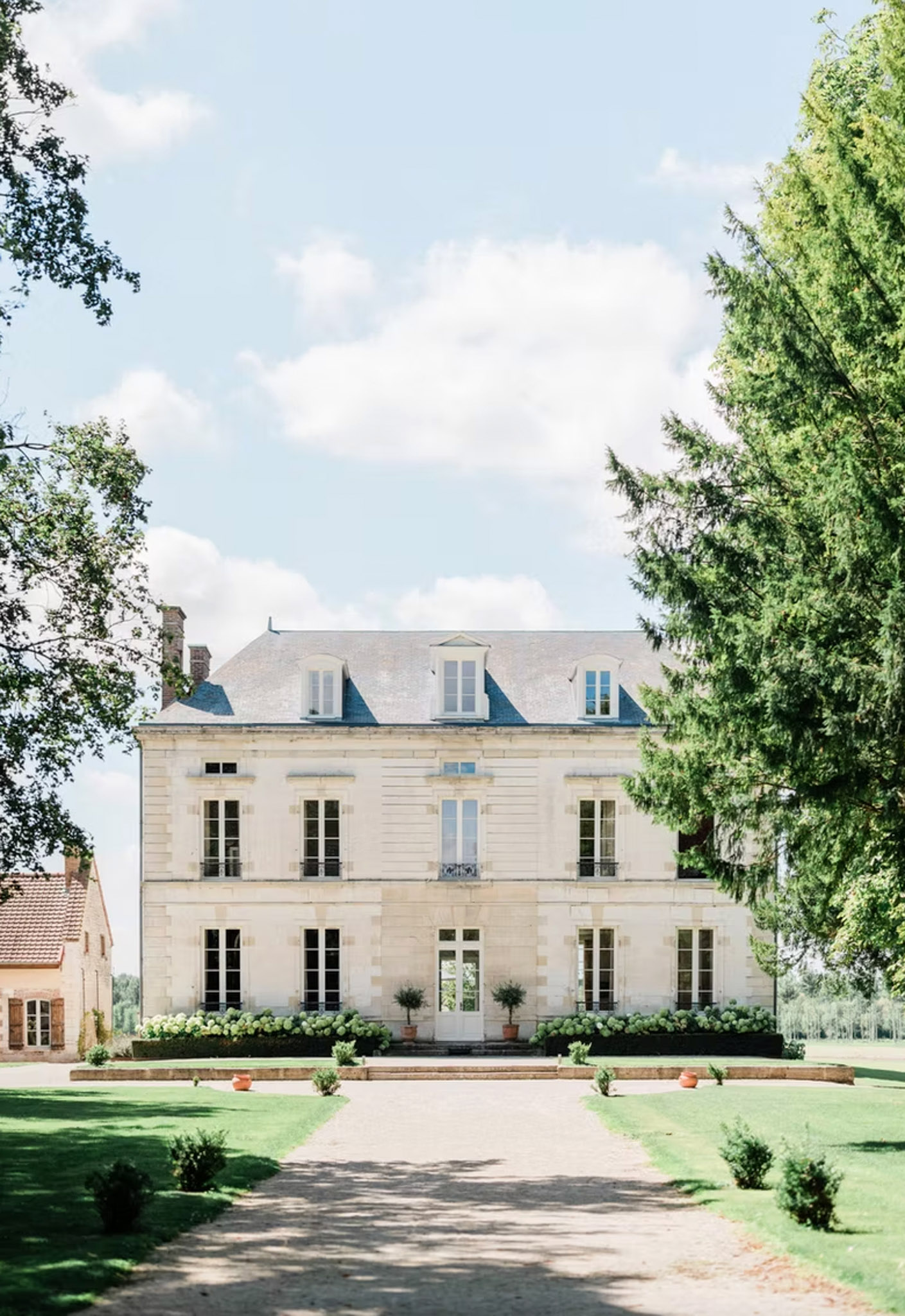 Cream stone French chateau with slate mansard roof, wrought iron balconies, white hydrangeas, and gravel driveway