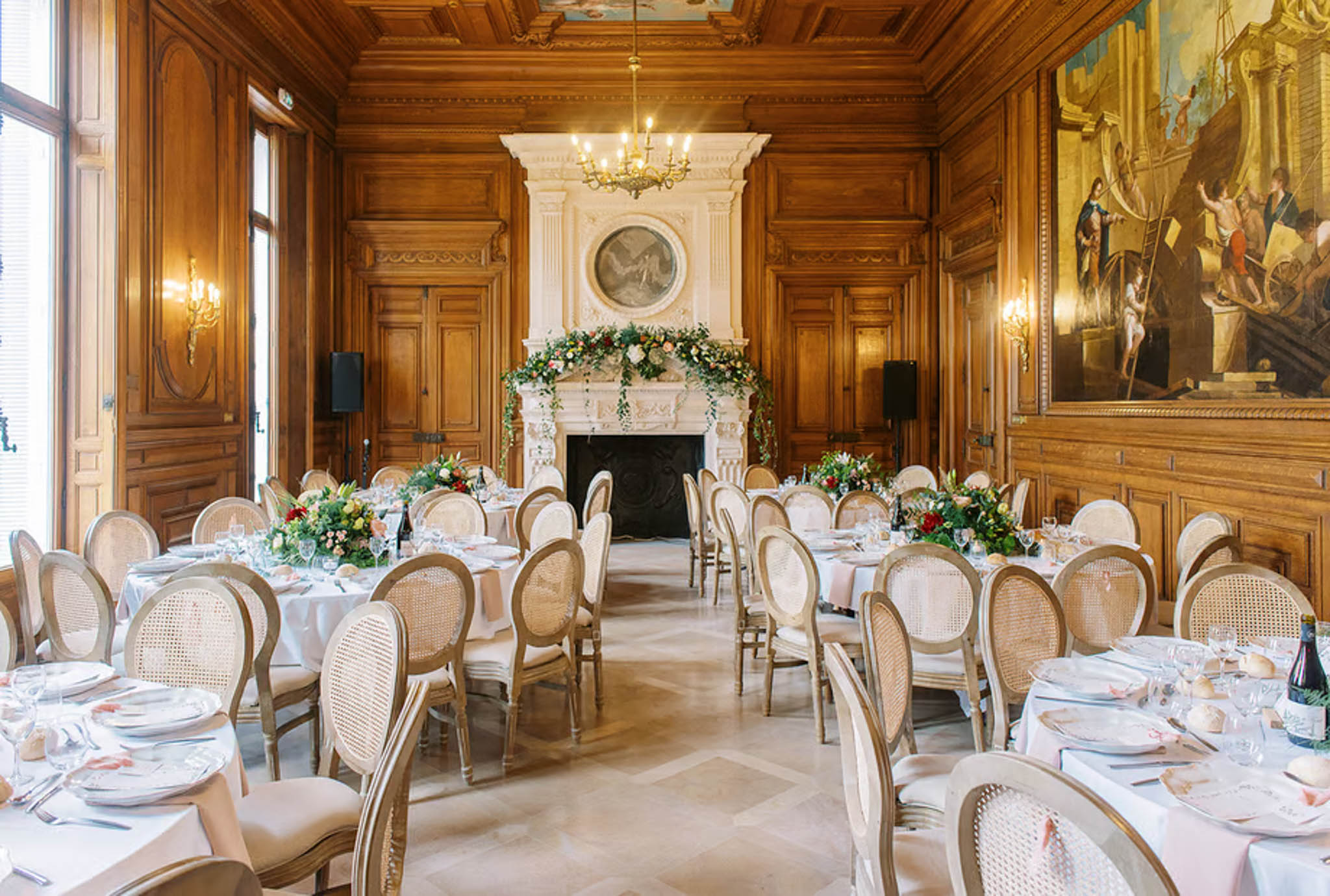 Wedding reception room in a French chateau with round tables, gold rattan chairs, and blush floral centerpieces by a stone...