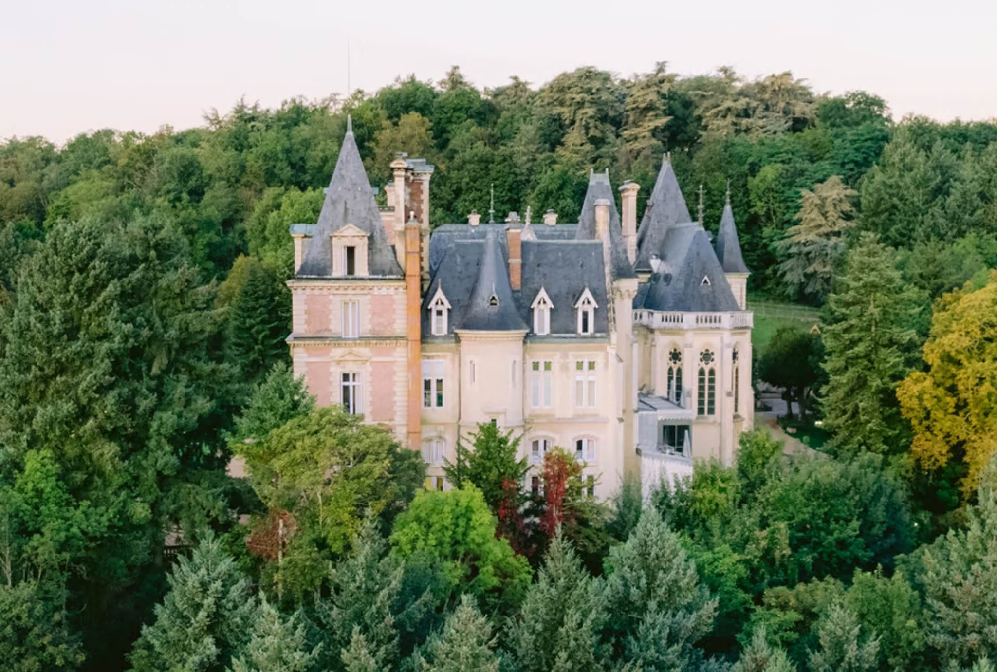 Aerial dusk view of a 19th-century French chateau with slate turrets and pink stone facades surrounded by woodland