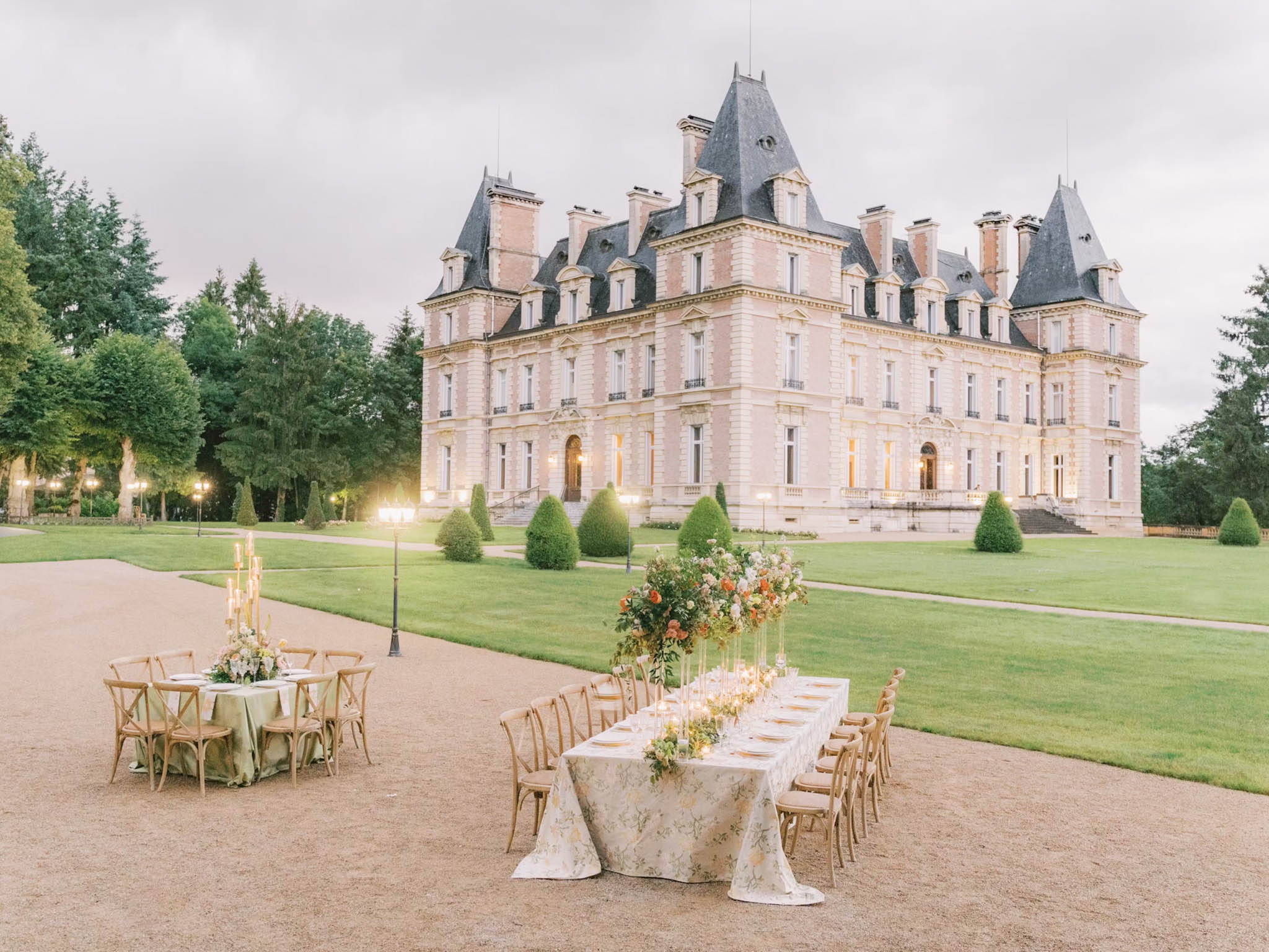 Outdoor reception tables with floral centerpieces and gold chargers on chateau gravel forecourt at dusk