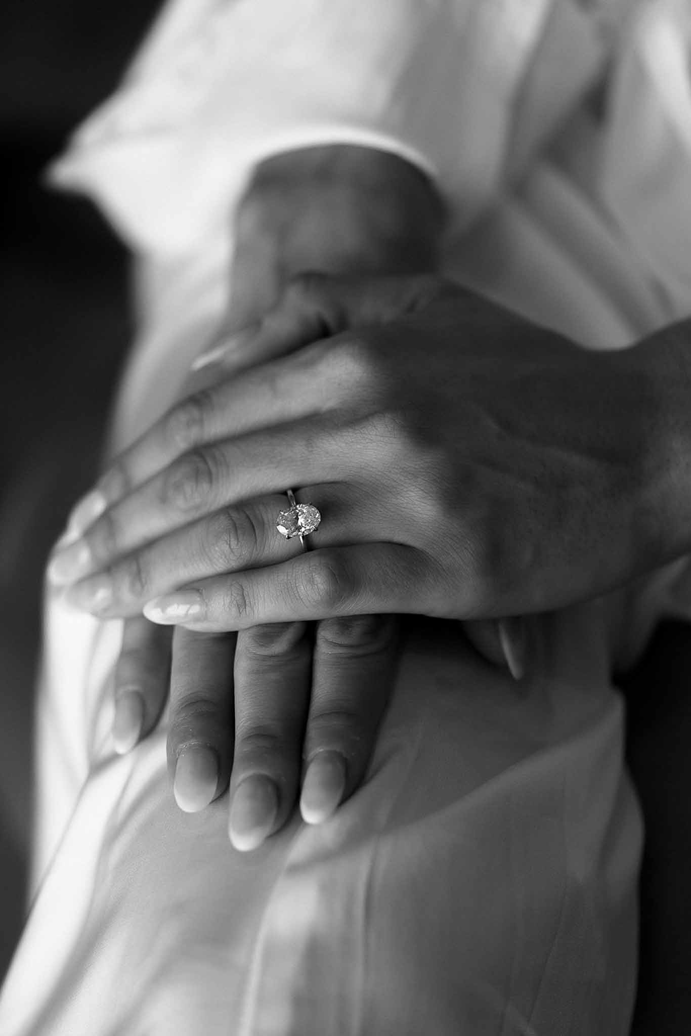 Black and white close-up of hands with oval solitaire engagement ring in intimate layered pose