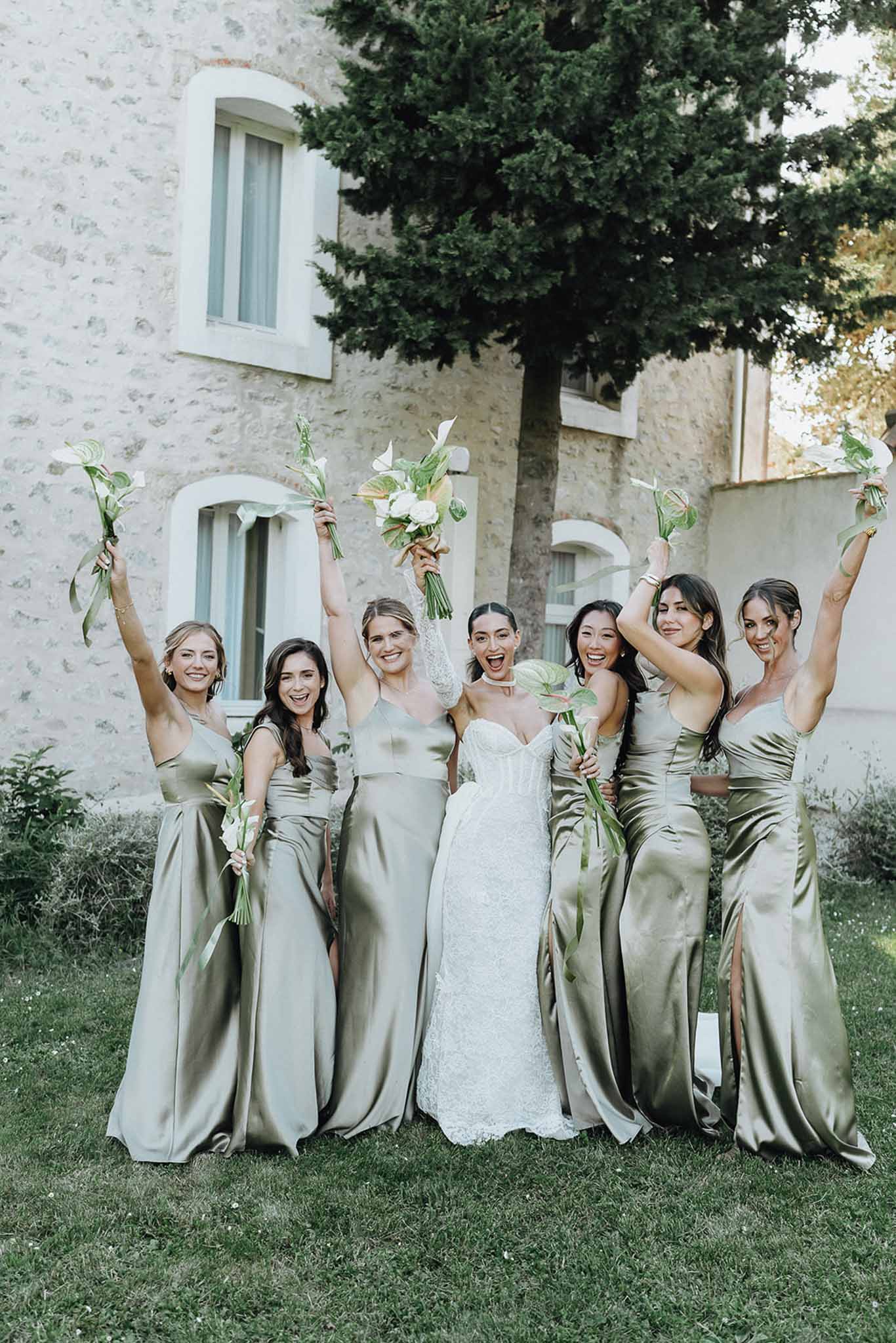 Bride in strapless lace gown with six bridesmaids in sage green satin dresses raising bouquets outside a French stone manor
