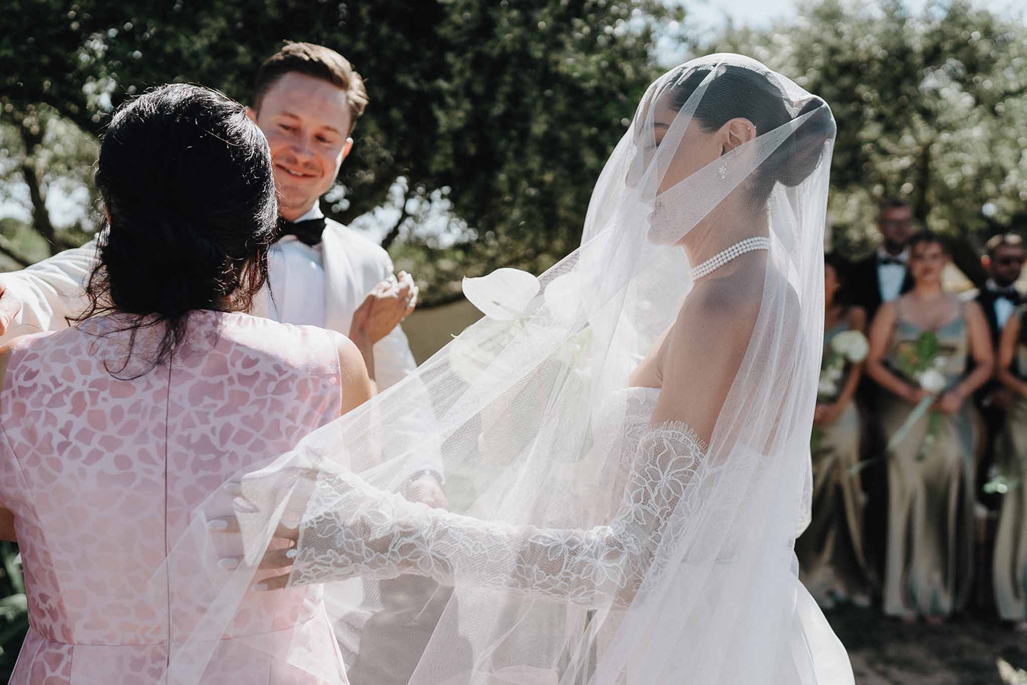 Woman lifting bride cathedral veil as groom in white suit jacket smiles during outdoor ceremony