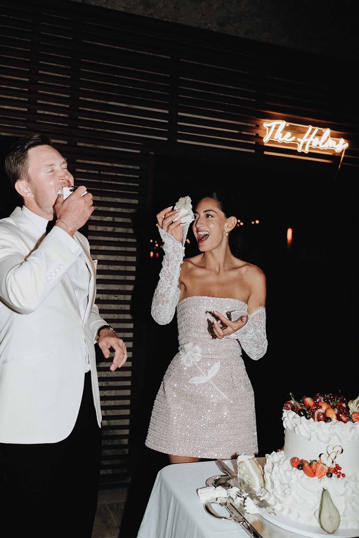 Bride and groom feeding each other cake beside fruit-topped white ruffled cake at night reception