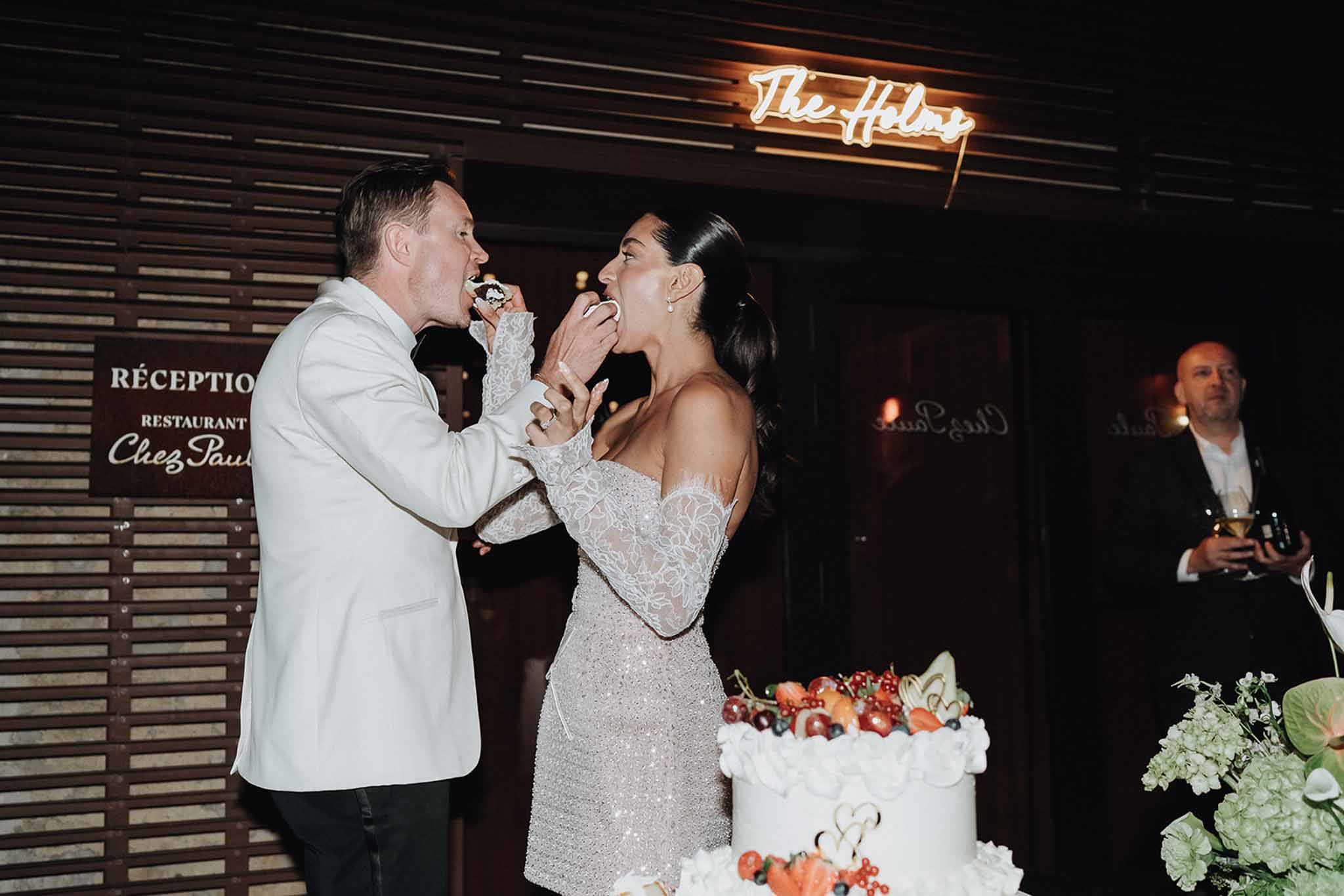 Couple feeding each other from ruffled two-tier cake with fresh berries and gold topper under amber neon sign