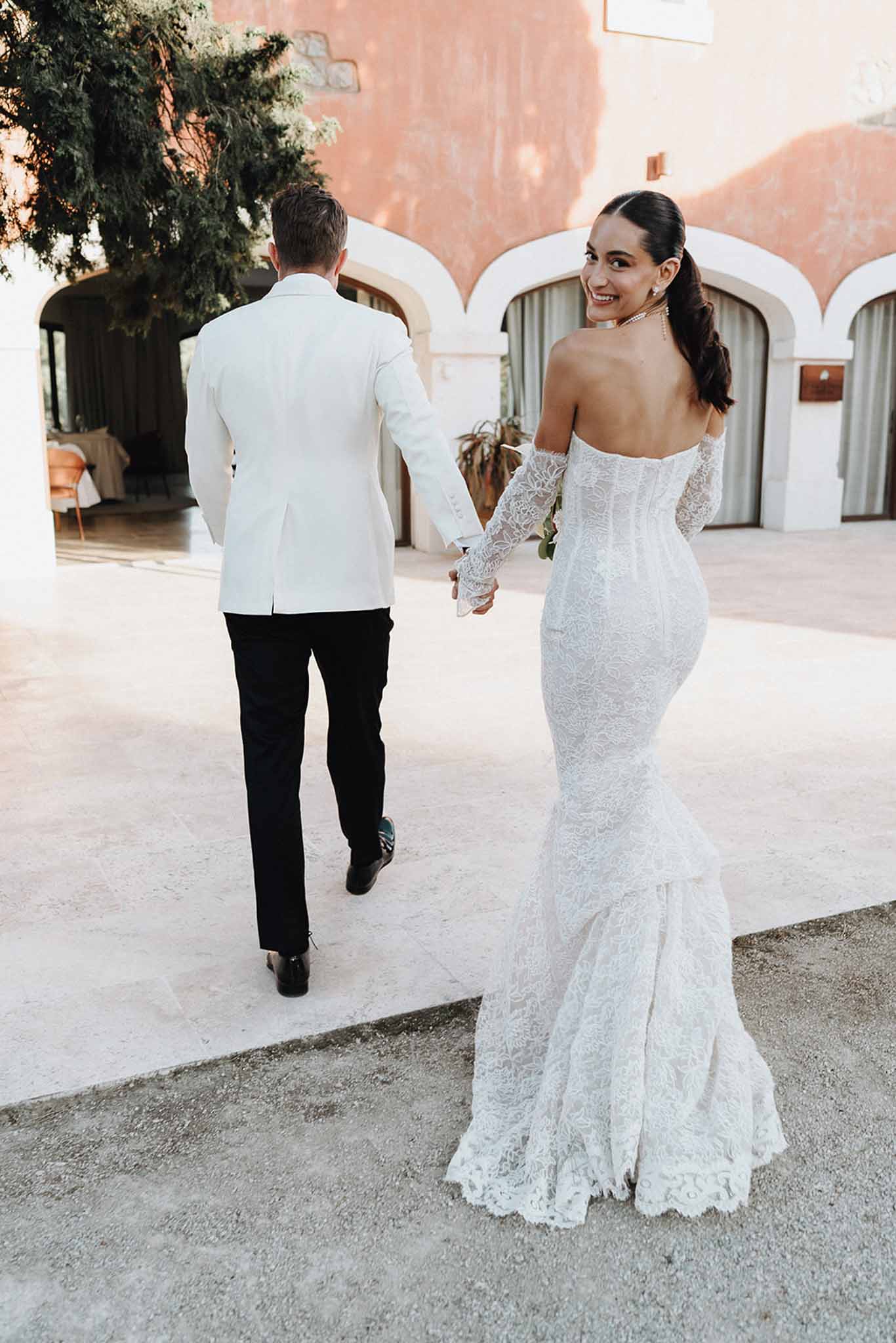 Couple walking away on stone courtyard with bride in off-shoulder lace mermaid gown glancing back