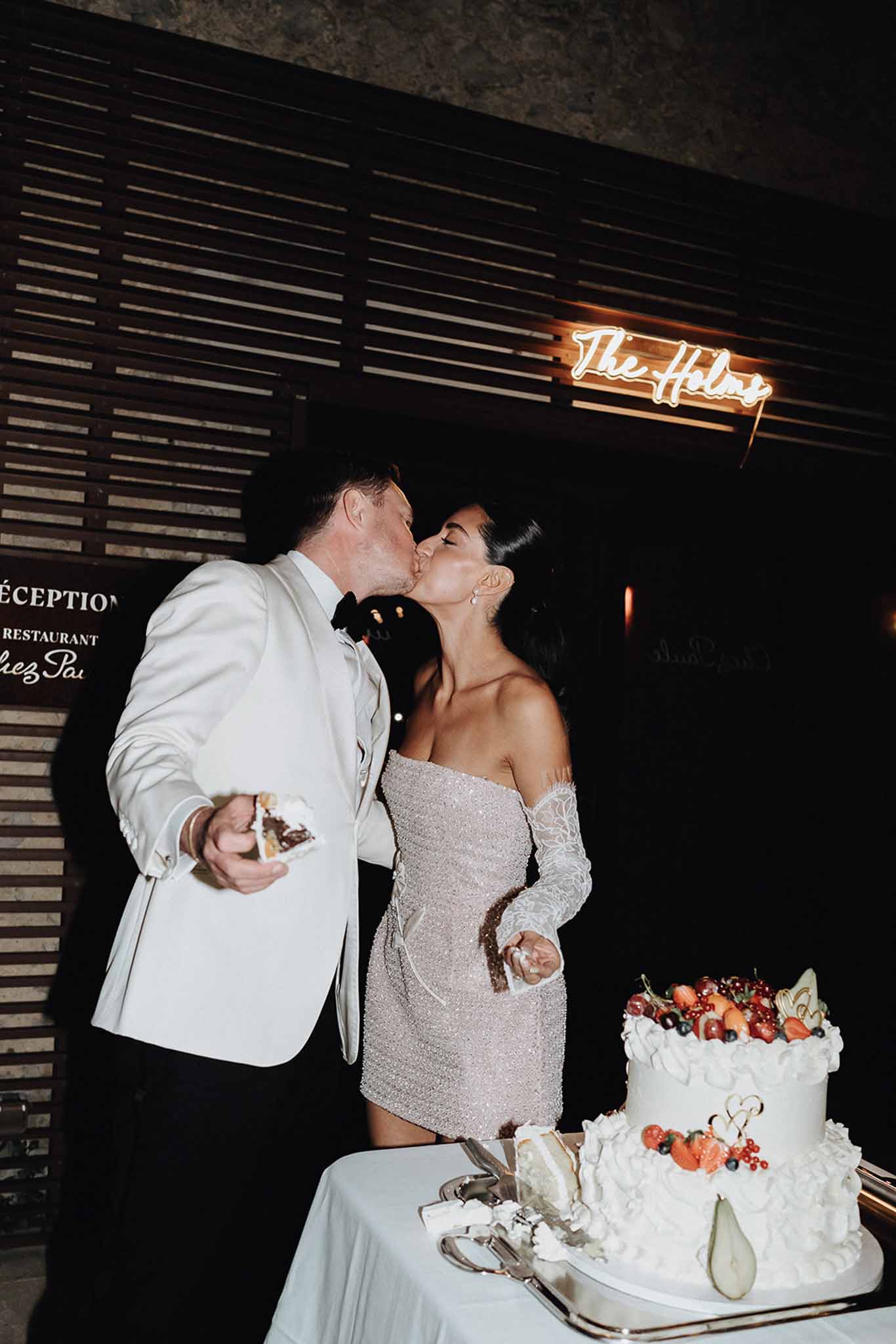 Couple kisses at cake cutting with two-tier berry-topped cake and orange neon sign in moody venue