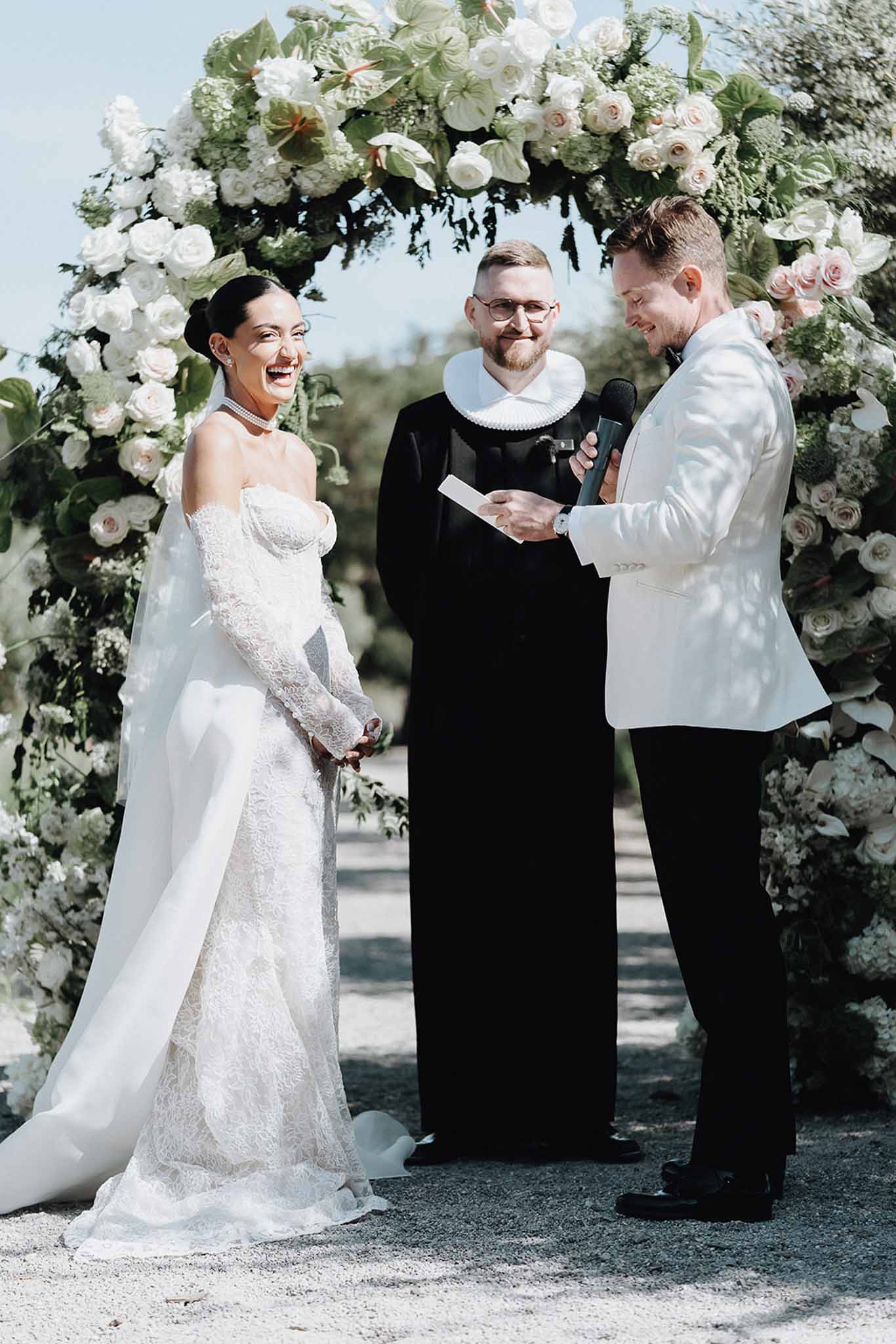 Laughing bride and groom exchanging vows under circular white rose and anthurium arch on gravel