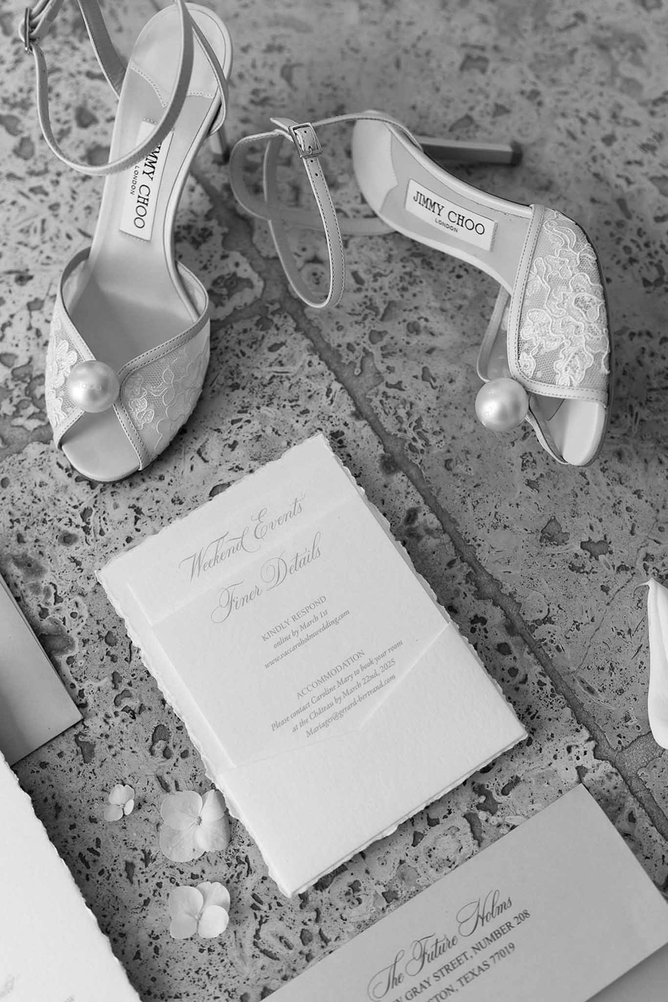 Black and white flat lay of Jimmy Choo lace heels with pearl toe beside weekend events card on stone surface
