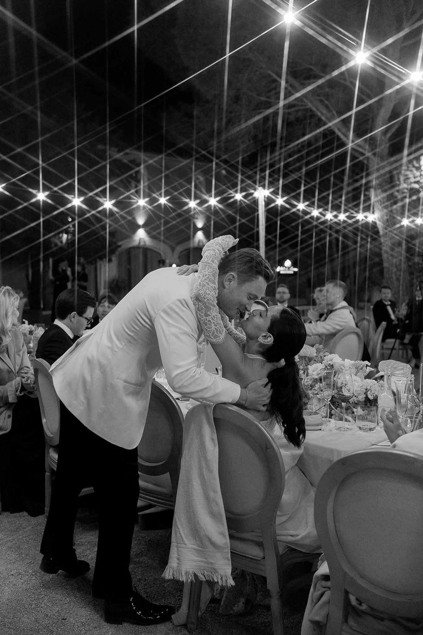 Black and white groom kissing seated bride at reception table under pendant lights in glass marquee