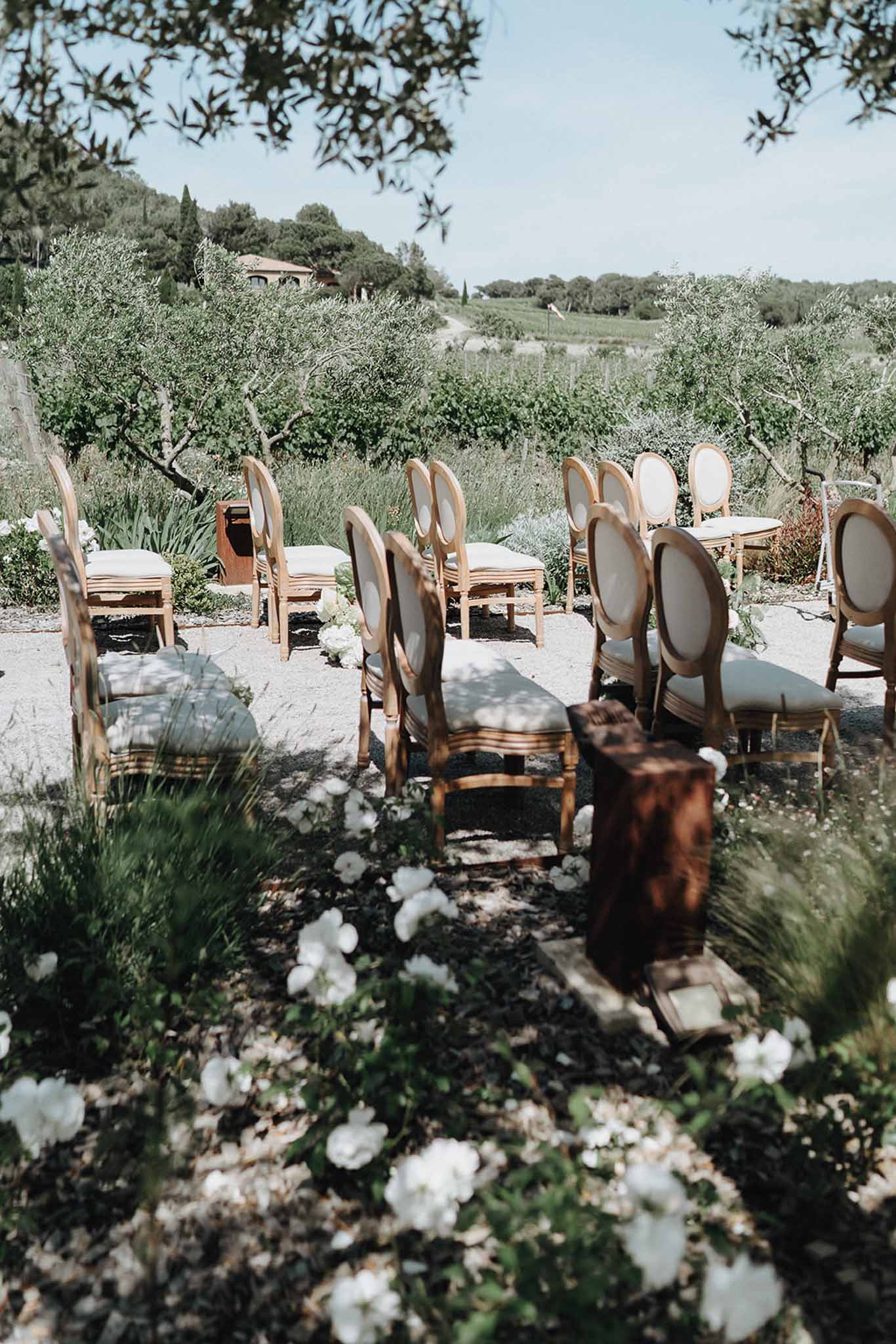 Louis XVI chairs on vineyard gravel with rustic lectern and white floral aisle clusters before stone farmhouse