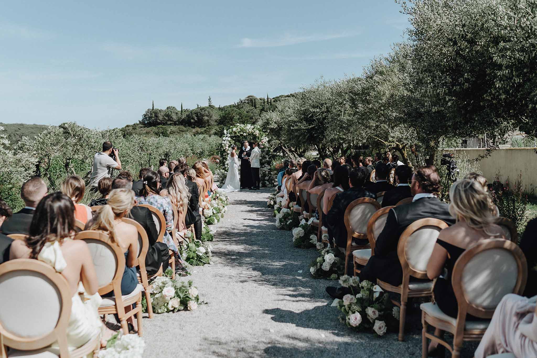 Couple at floral arch altar in olive grove as 70 guests sit in gold chairs with white rose aisle markers