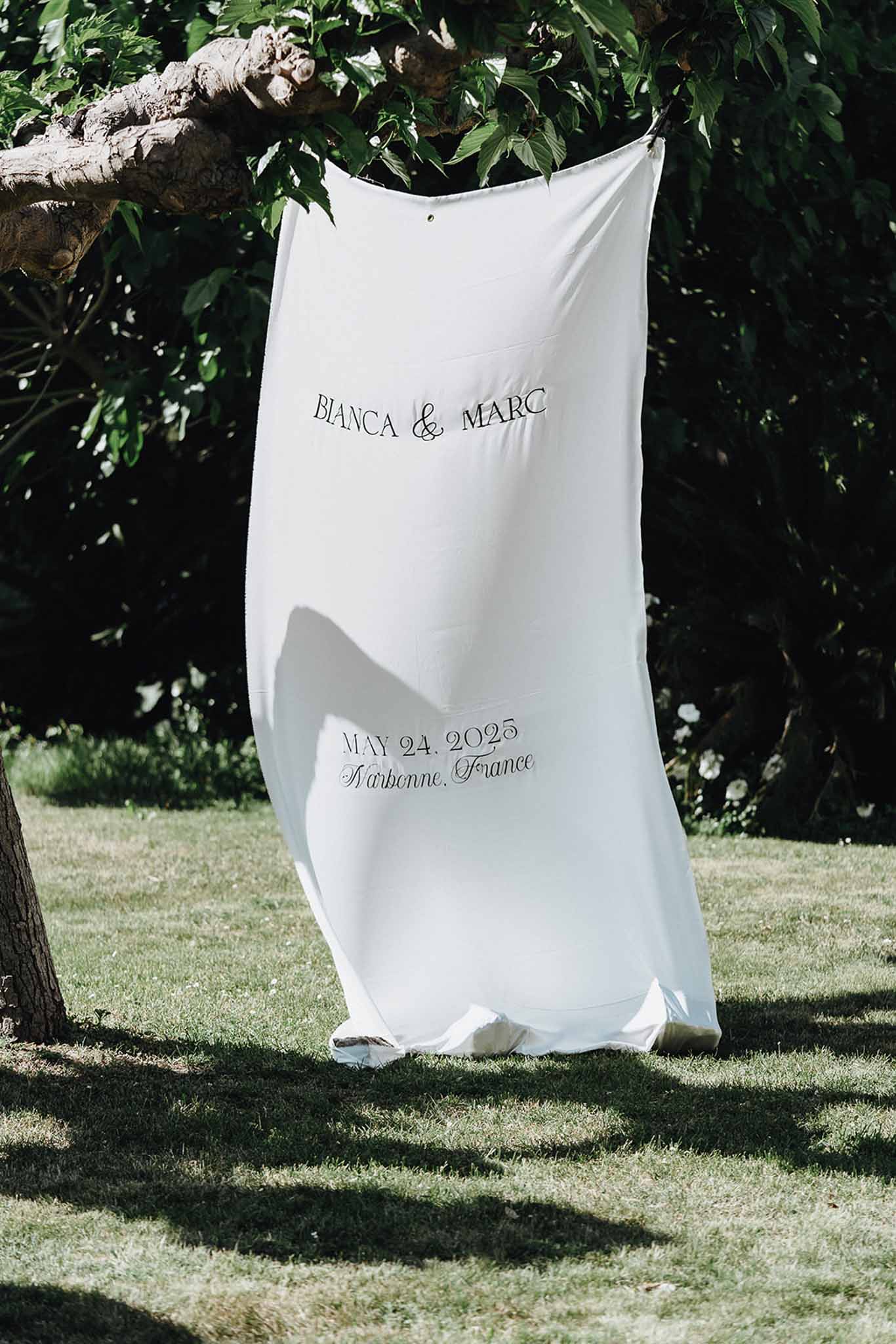White fabric wedding banner with couple names and date hanging from tree branch in garden