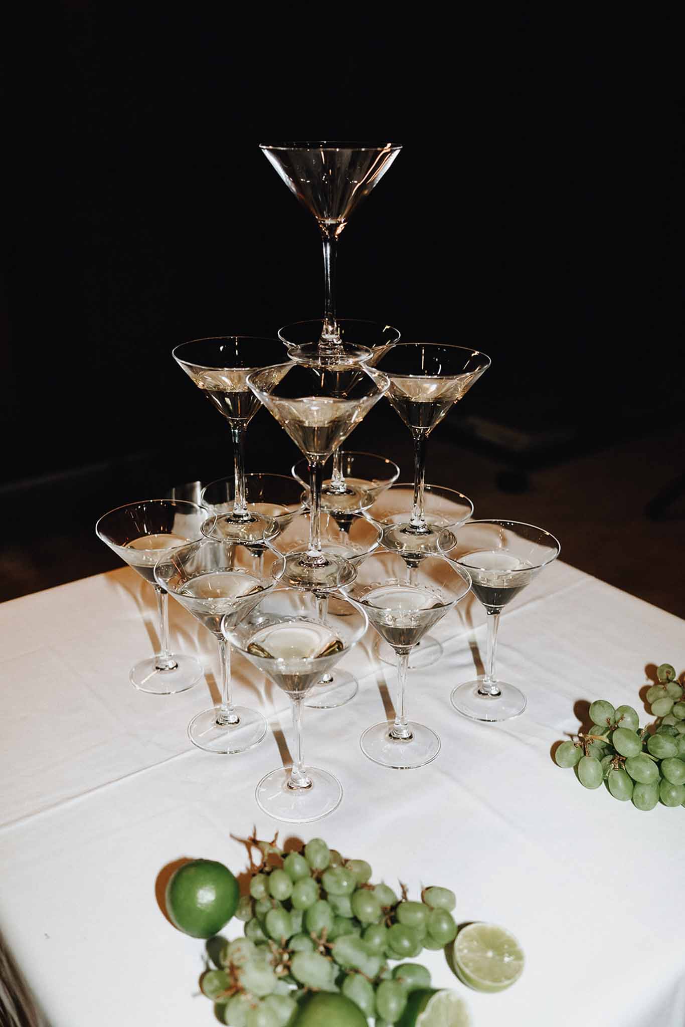 Martini glass pyramid tower on white linen with grape and lime garnish against dark background at reception