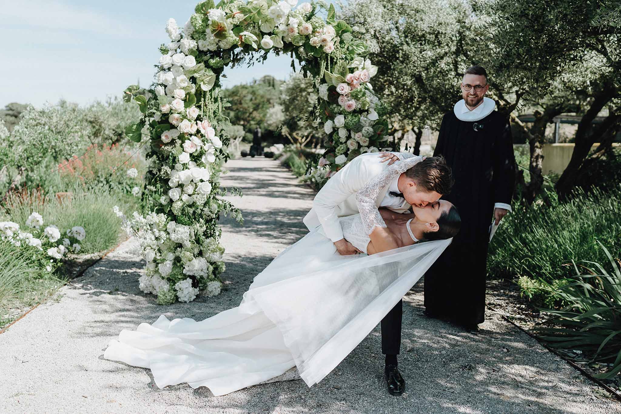 Dip kiss under circular white rose and anthurium arch on olive tree-lined gravel path