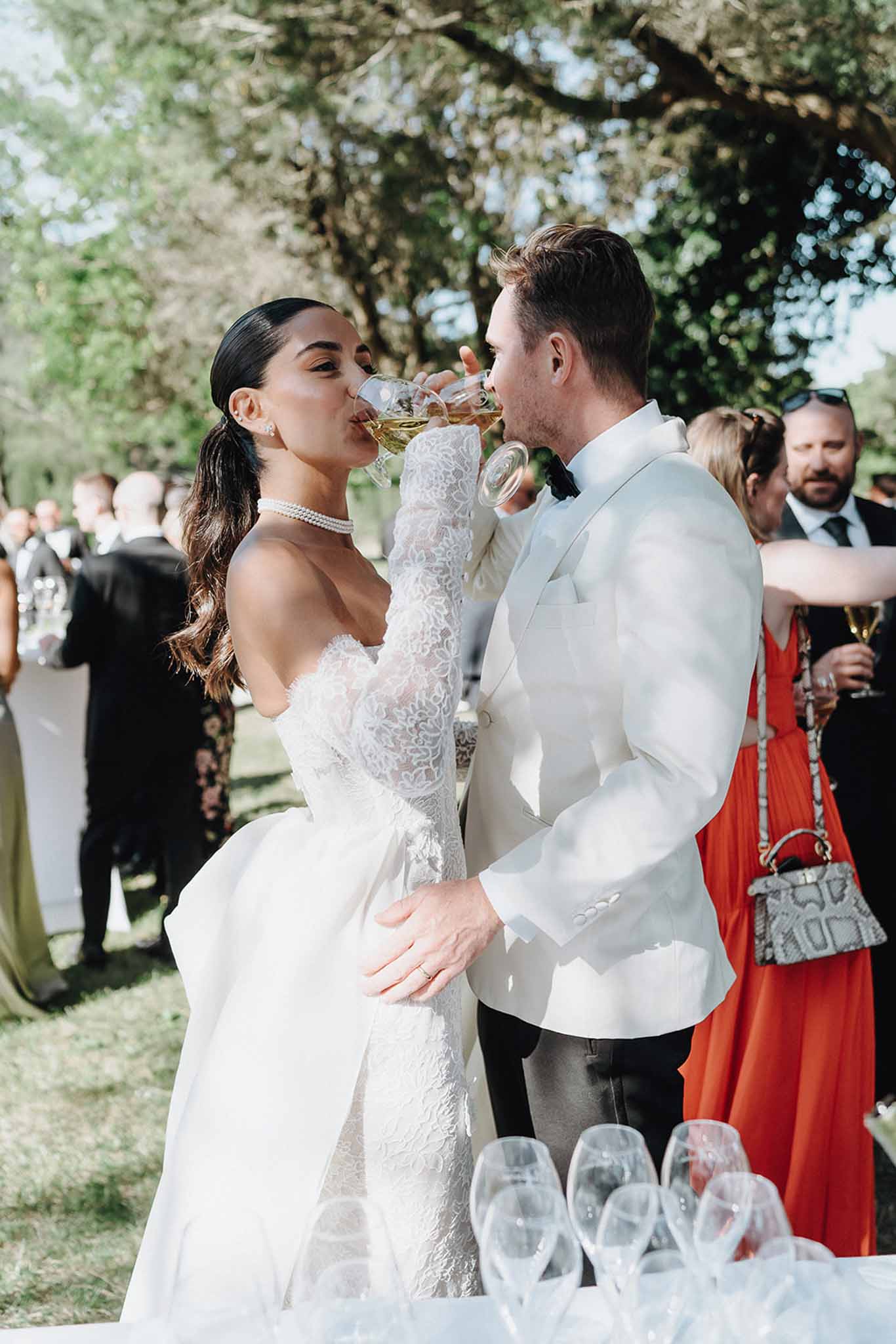 Couple crossing champagne flutes in toast, bride in lace gown with pearl choker, groom in white dinner jacket