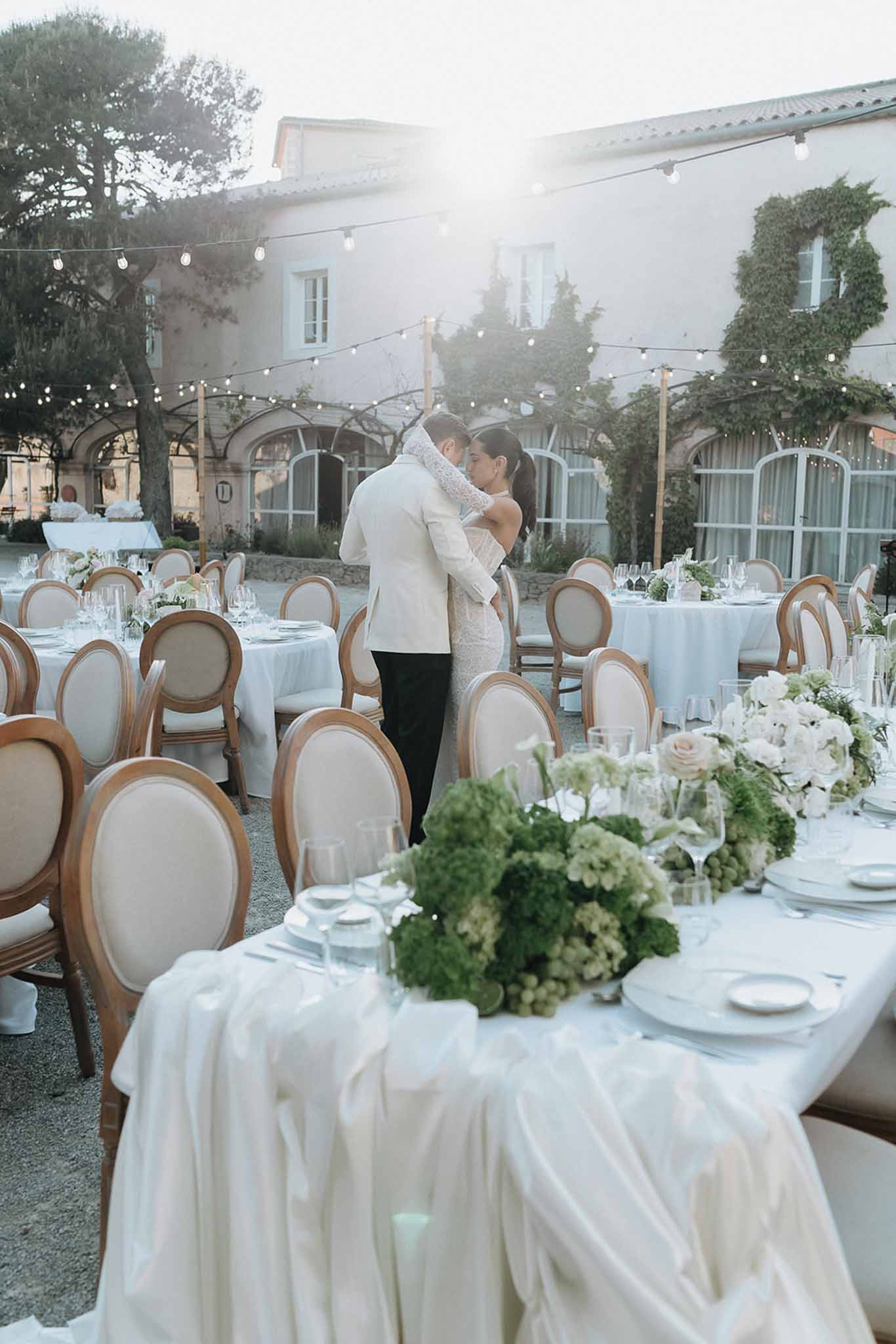 Couple embraces at dusk among round tables with green hydrangea centerpieces and globe lights at domaine