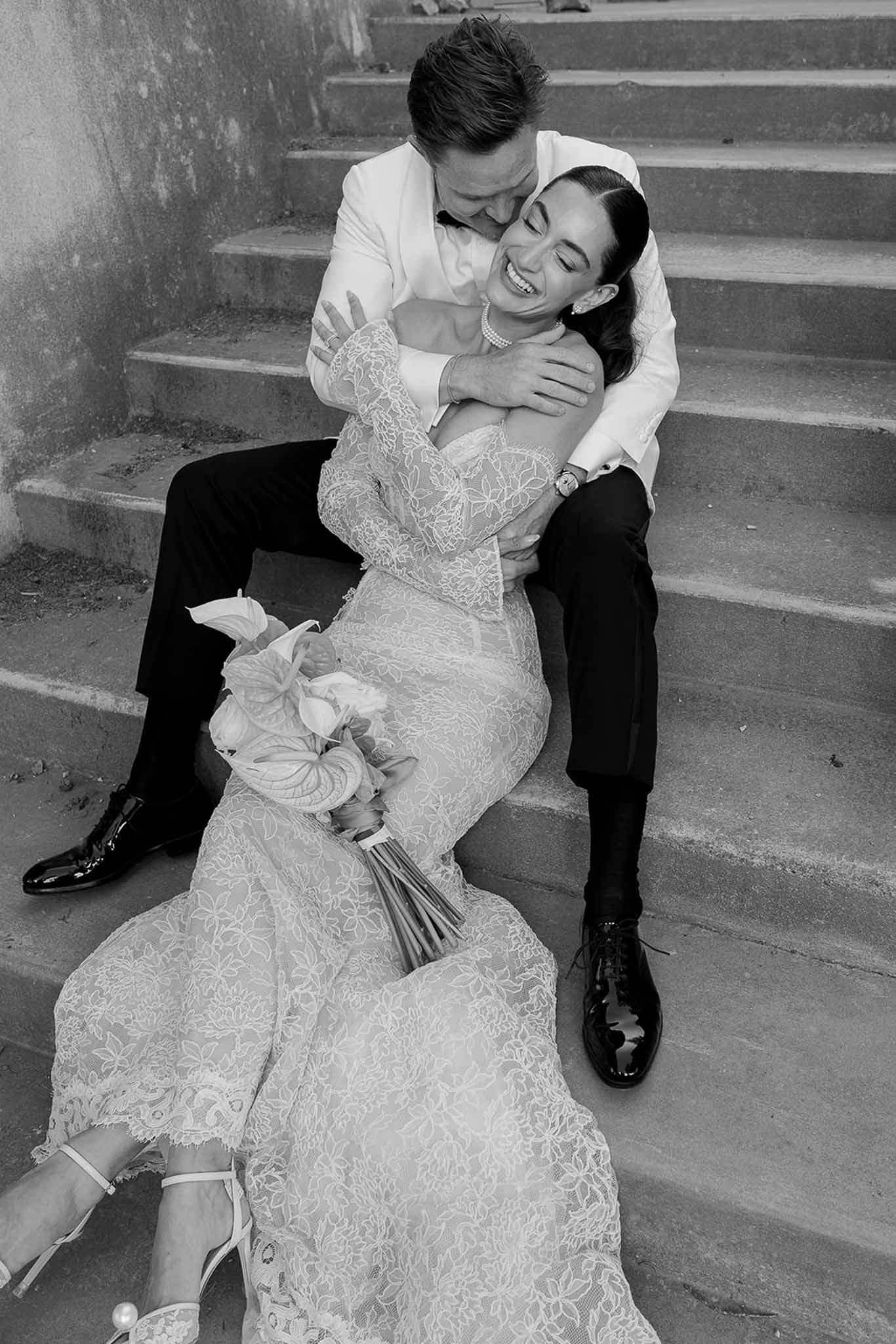 Black-and-white portrait of bride and groom laughing on stone chateau staircase in lace gown and white jacket