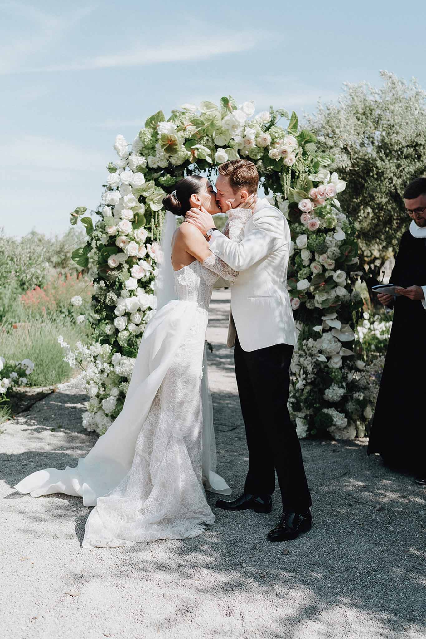 First kiss under circular arch of white roses, blush roses, and green anthurium on gravel