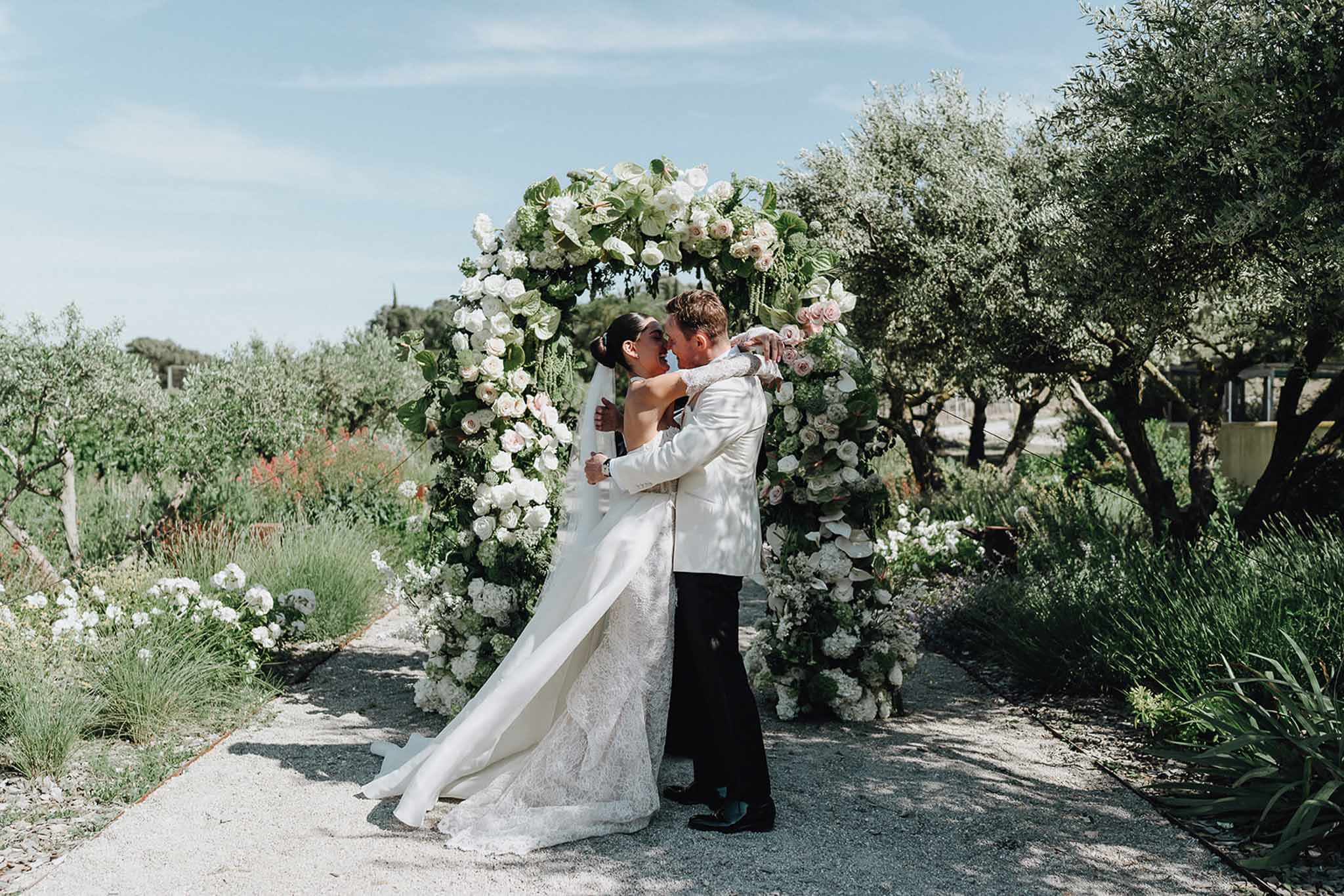 Bride and groom kissing in front of circular floral arch of white and blush roses in a Mediterranean garden setting