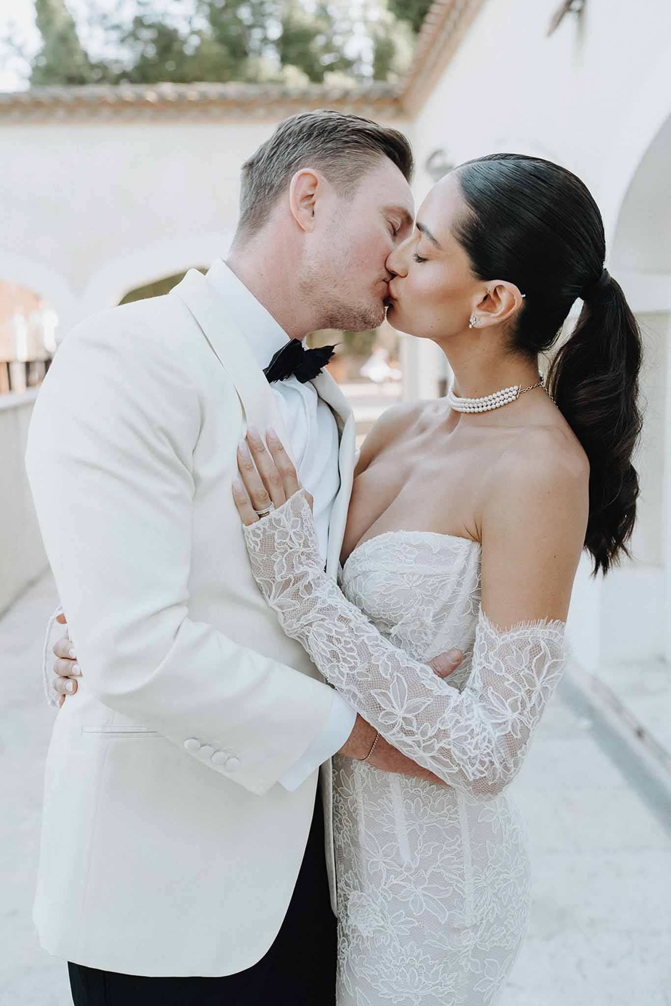 Bride and groom kissing outdoors, bride in off-the-shoulder lace gown with pearl choker, groom in white dinner jacket