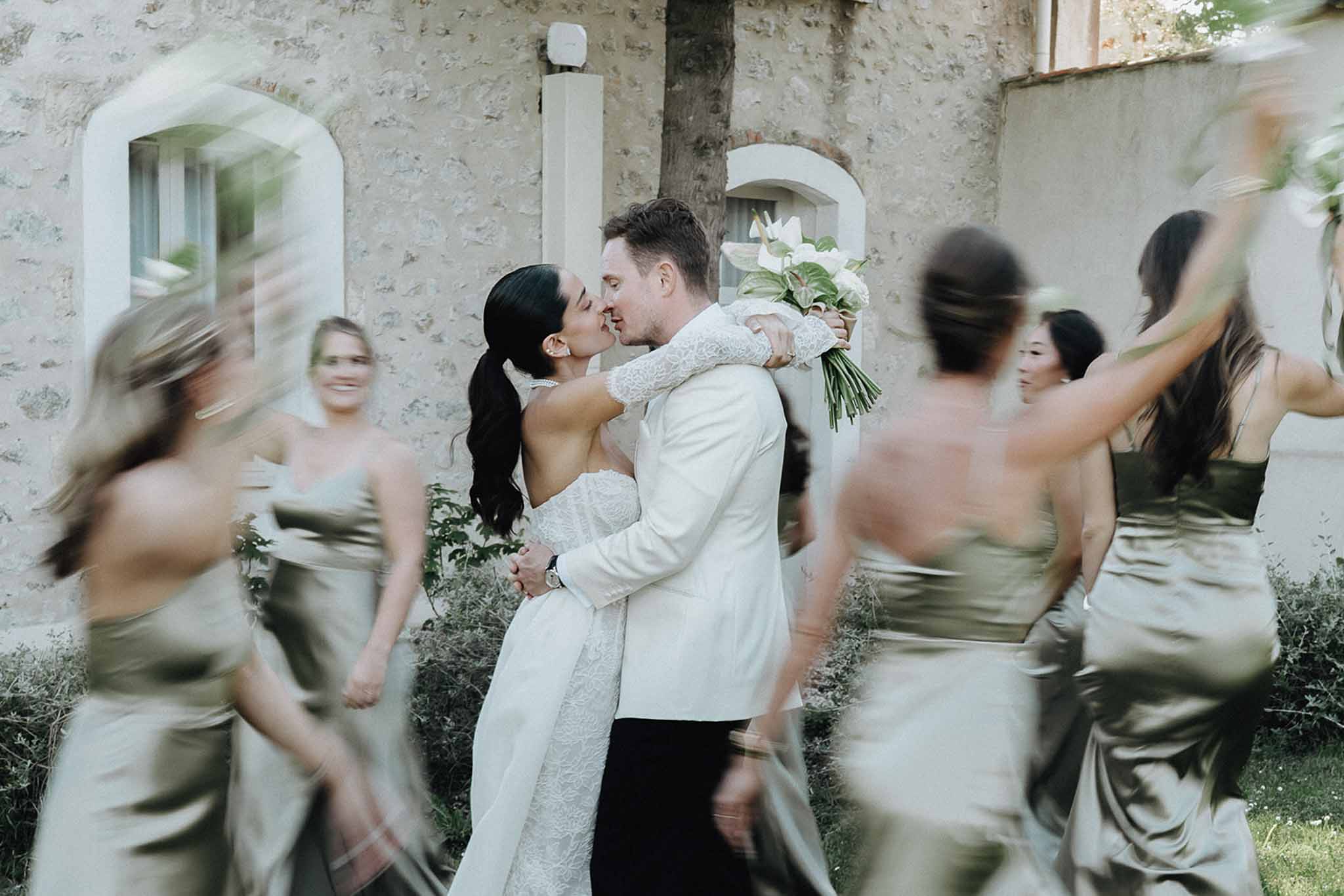 Couple kissing sharp while five sage green bridesmaids circle in motion blur before stone chateau