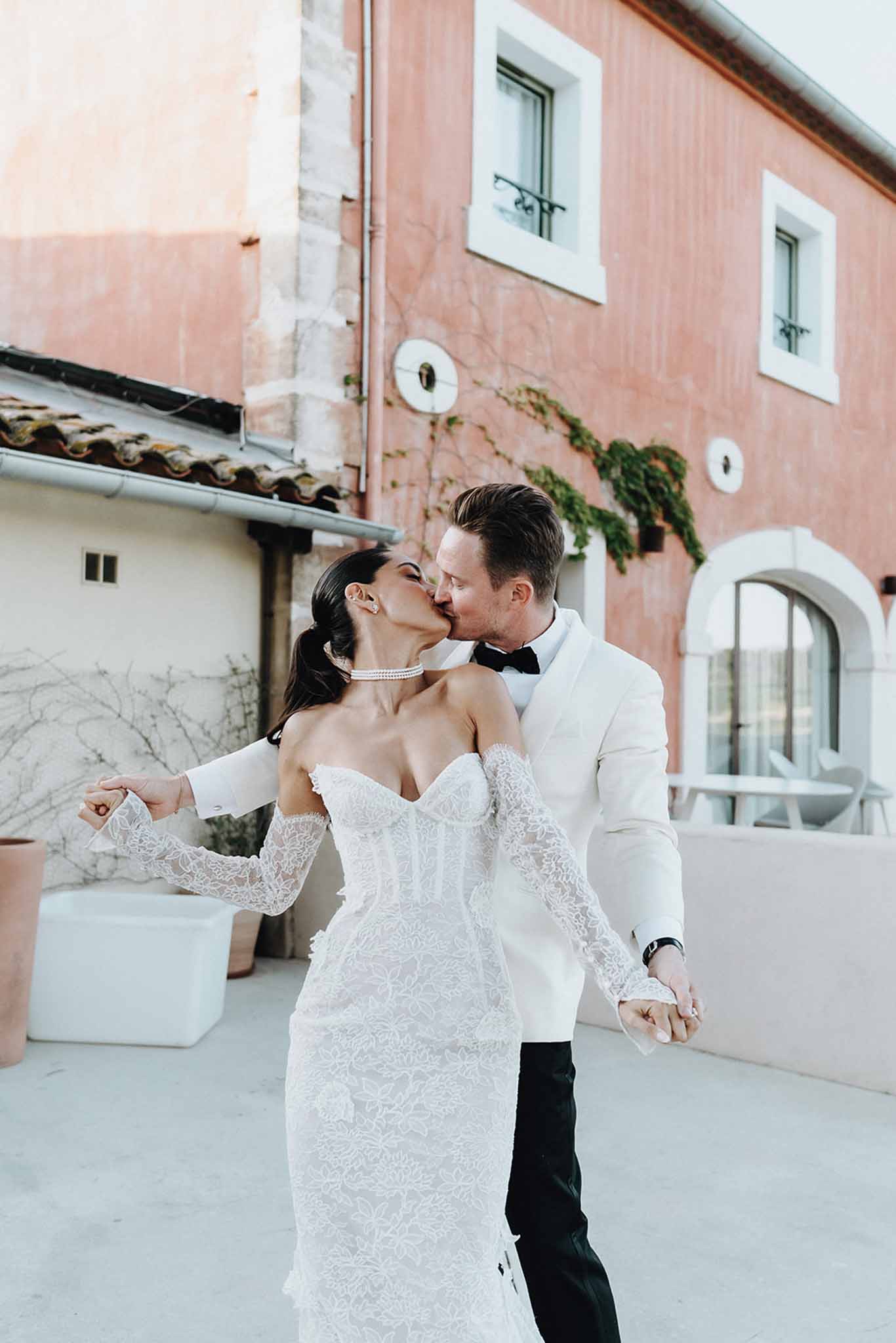 Couple kissing on terrace before terracotta-pink Provencal building, bride in lace gown with pearl choker necklace