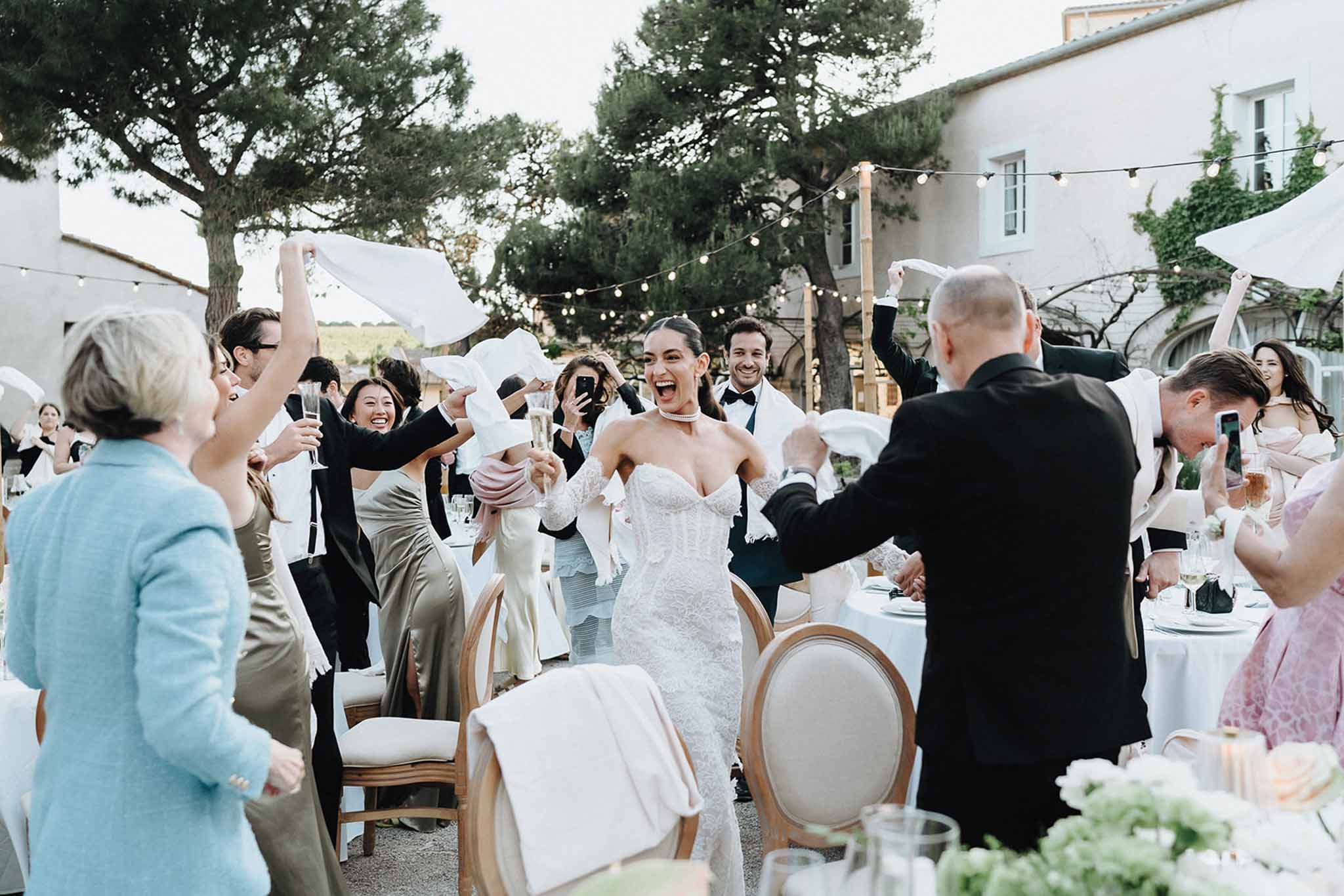 Bride in lace gown with champagne flute entering reception as 25 guests wave white napkins overhead